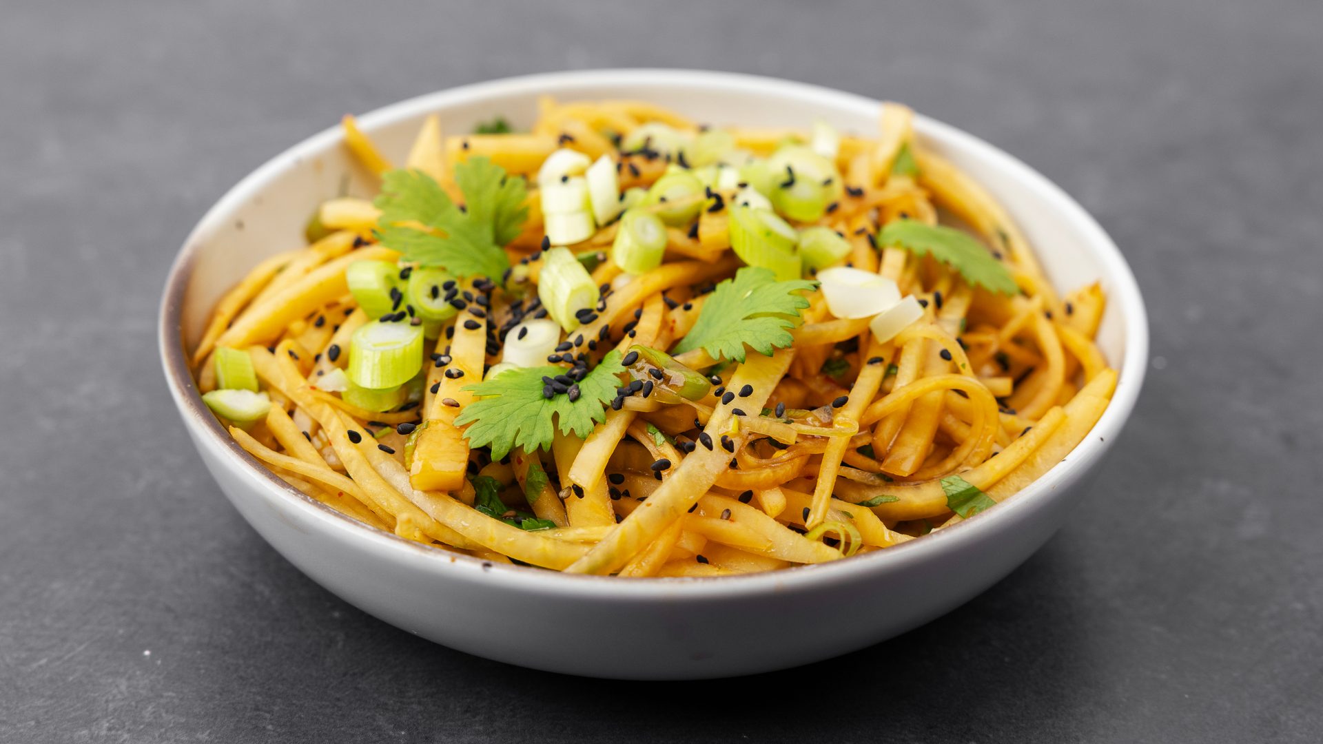 A bowl of yellow noodles or mango salad topped with green onions, cilantro, and black sesame seeds.