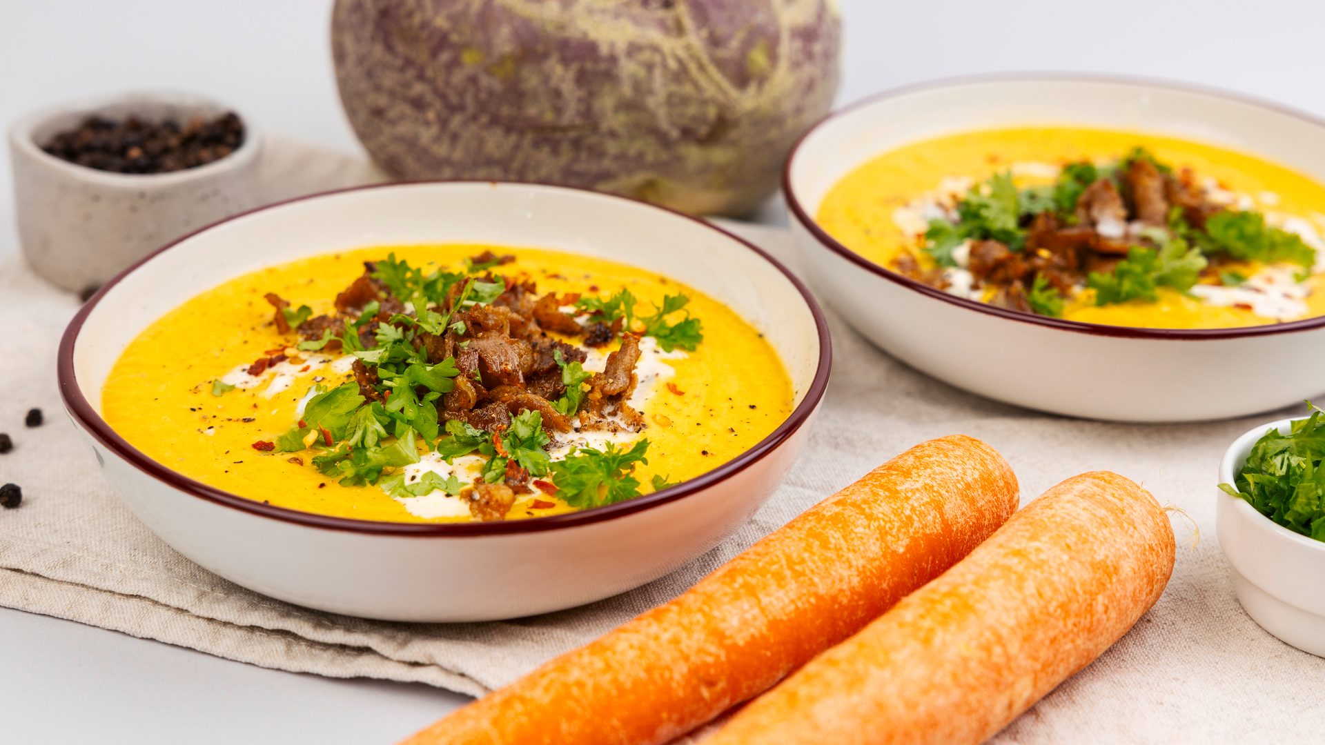 Two bowls of vibrant yellow soup, garnished with fresh parsley, cream, and crispy meat bits, with carrots.