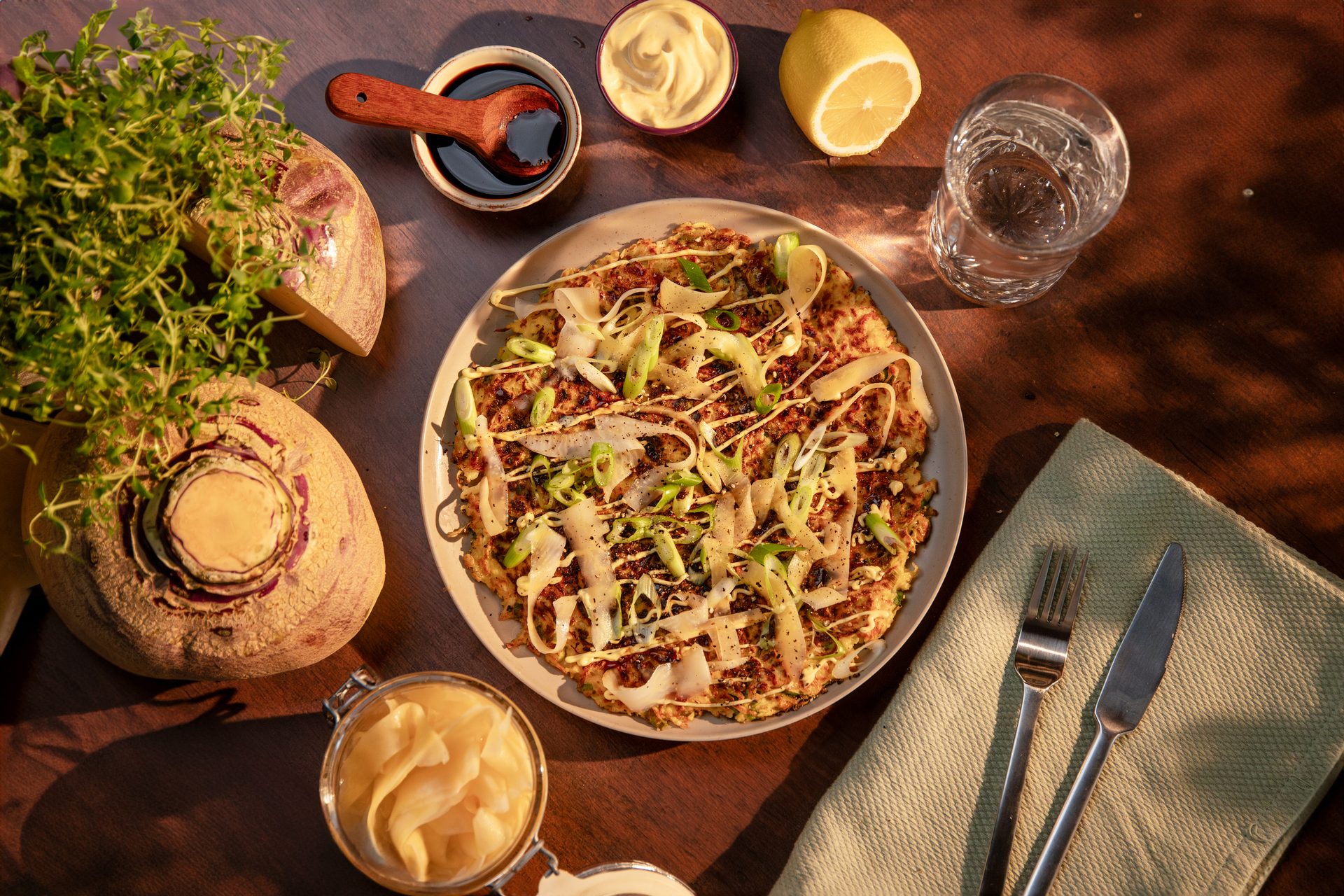 A savory pancake (Okonomiyaki) with toppings, surrounded by fresh kohlrabis, sauces, lemon, and water.