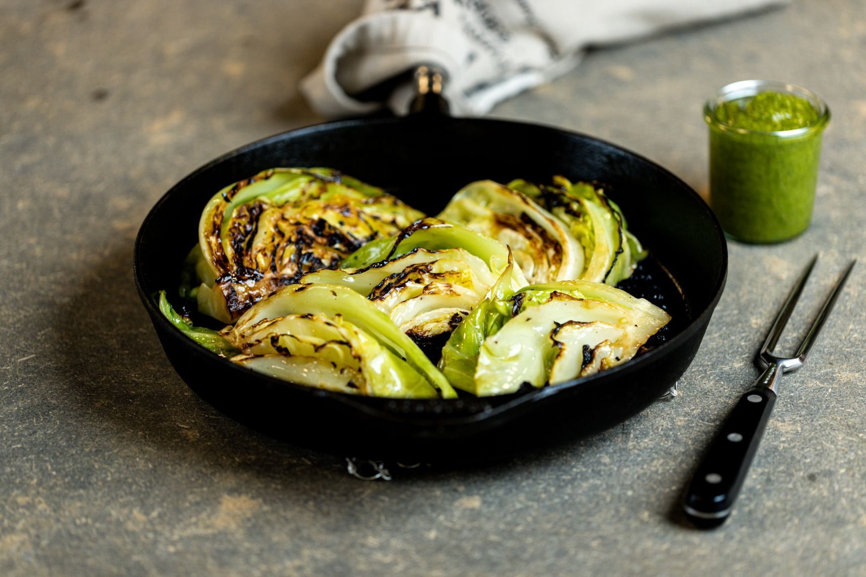 Grilled cabbage in a cast iron pan, with a jar of green sauce and a carving fork on the side.