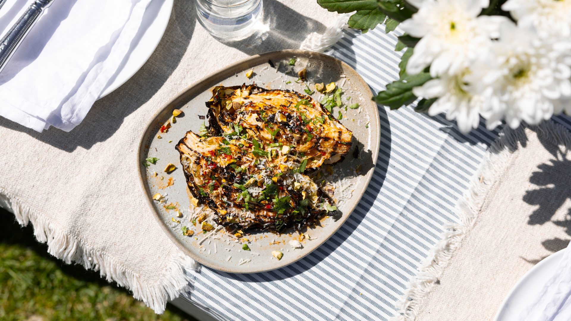 Grilled cabbage steaks garnished with herbs and nuts, served on a plate outdoors with a festive table setting.