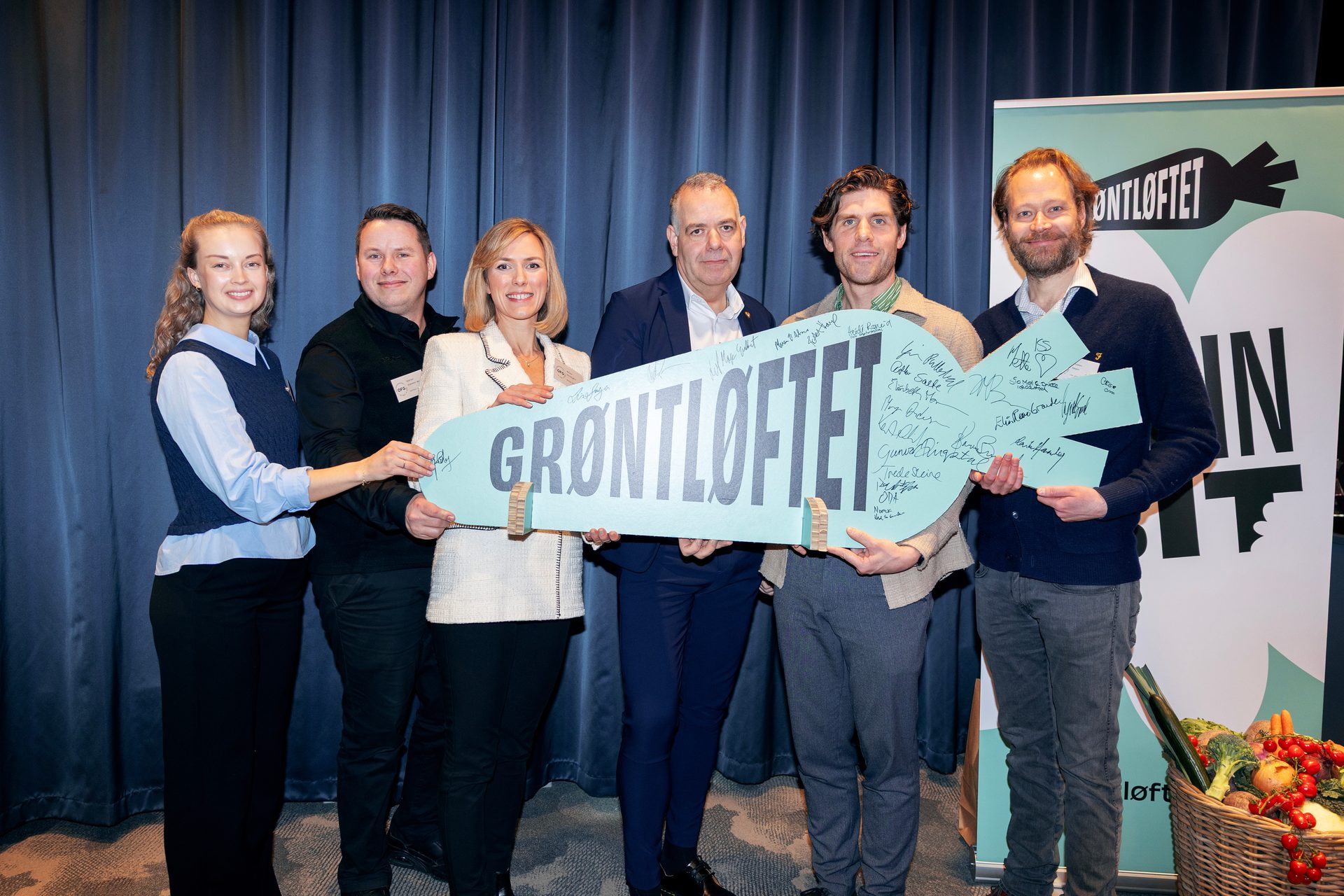 Six people hold a large key-shaped "GRØNTLØFTET" sign at an event with a banner.