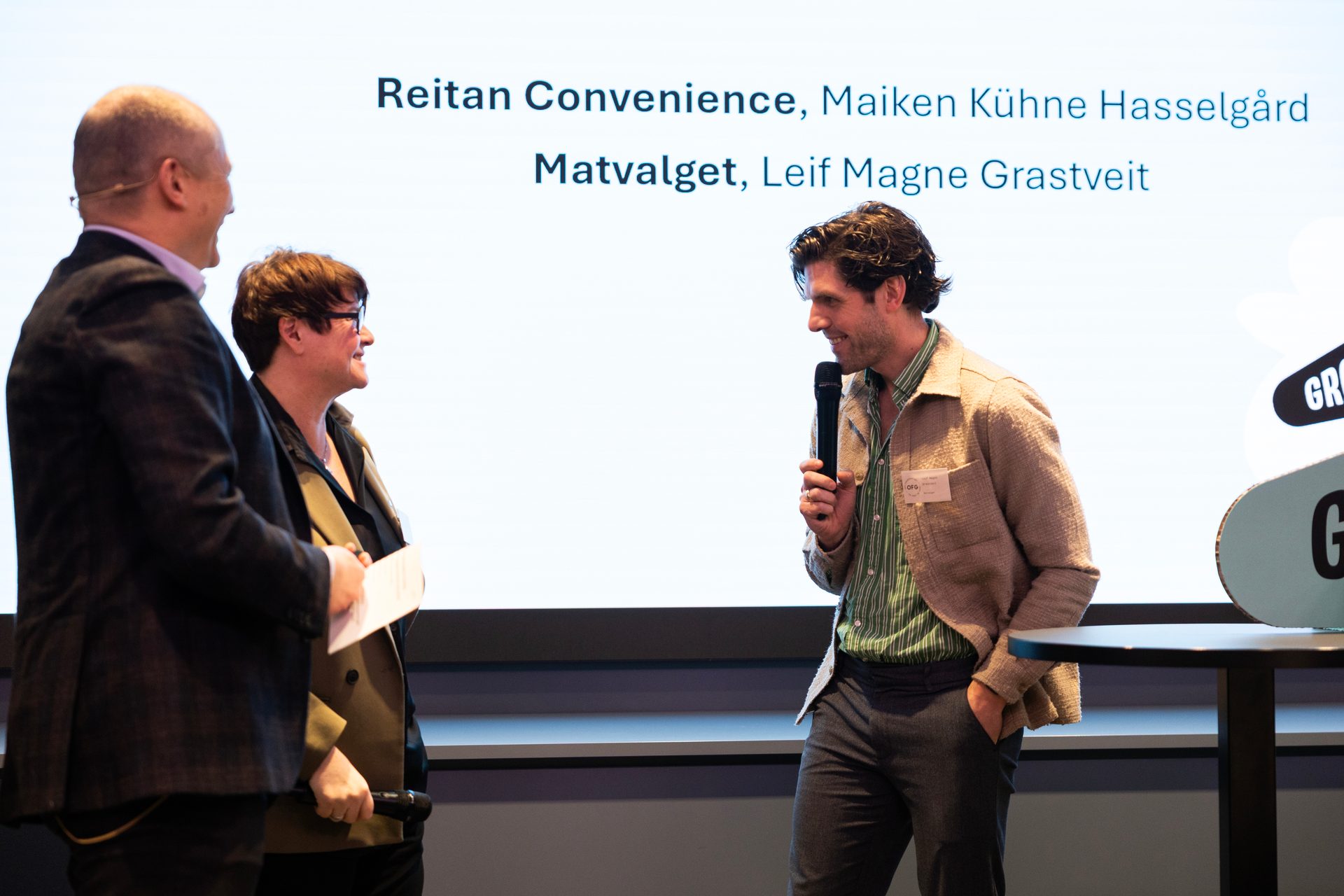 Three people on stage during a presentation, one speaking into a mic, another holding papers.