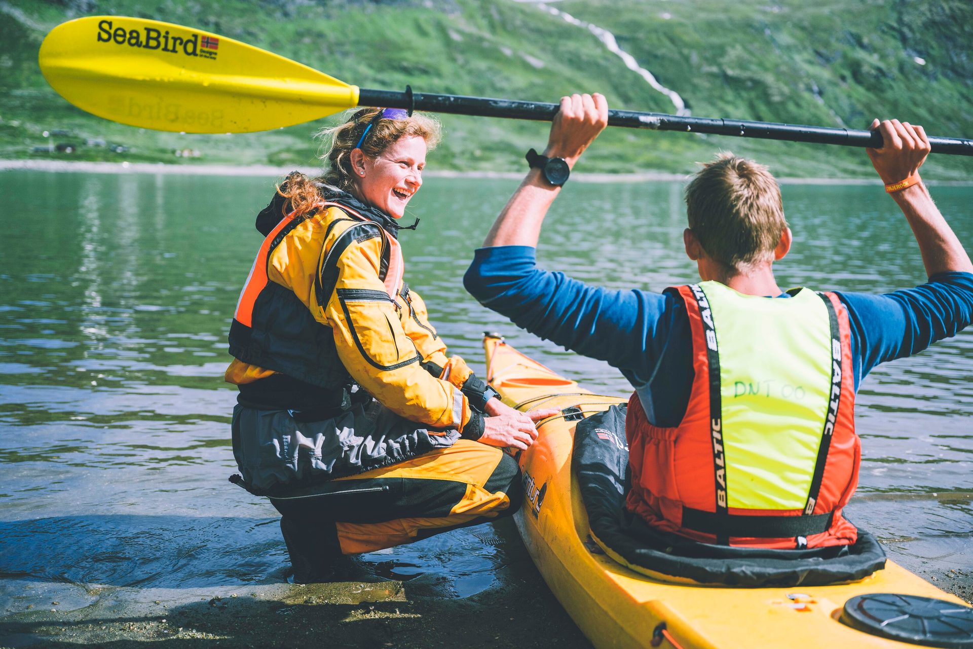 Two happy people with kayaks by a lake; one laughing, the other seated with a paddle.