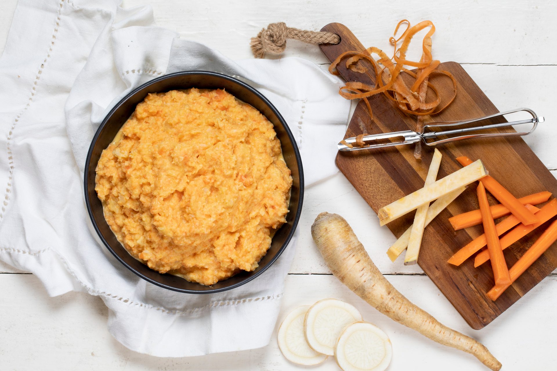 A bowl of orange root vegetable mash with carrots, parsnips, and a peeler on a white wooden surface.