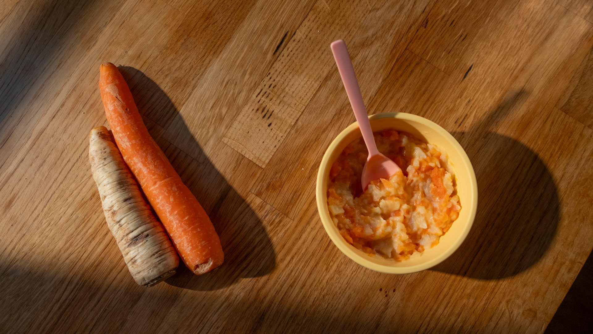 Two carrots and a bowl of baby food with a spoon on a wooden table.