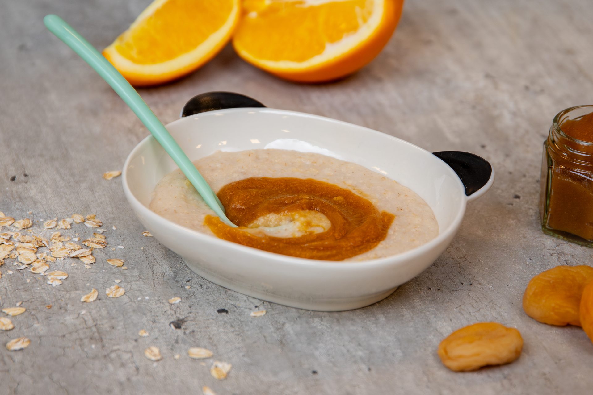 A white bowl of creamy oatmeal topped with a swirl of orange puree, accompanied by orange slices and a spoon.