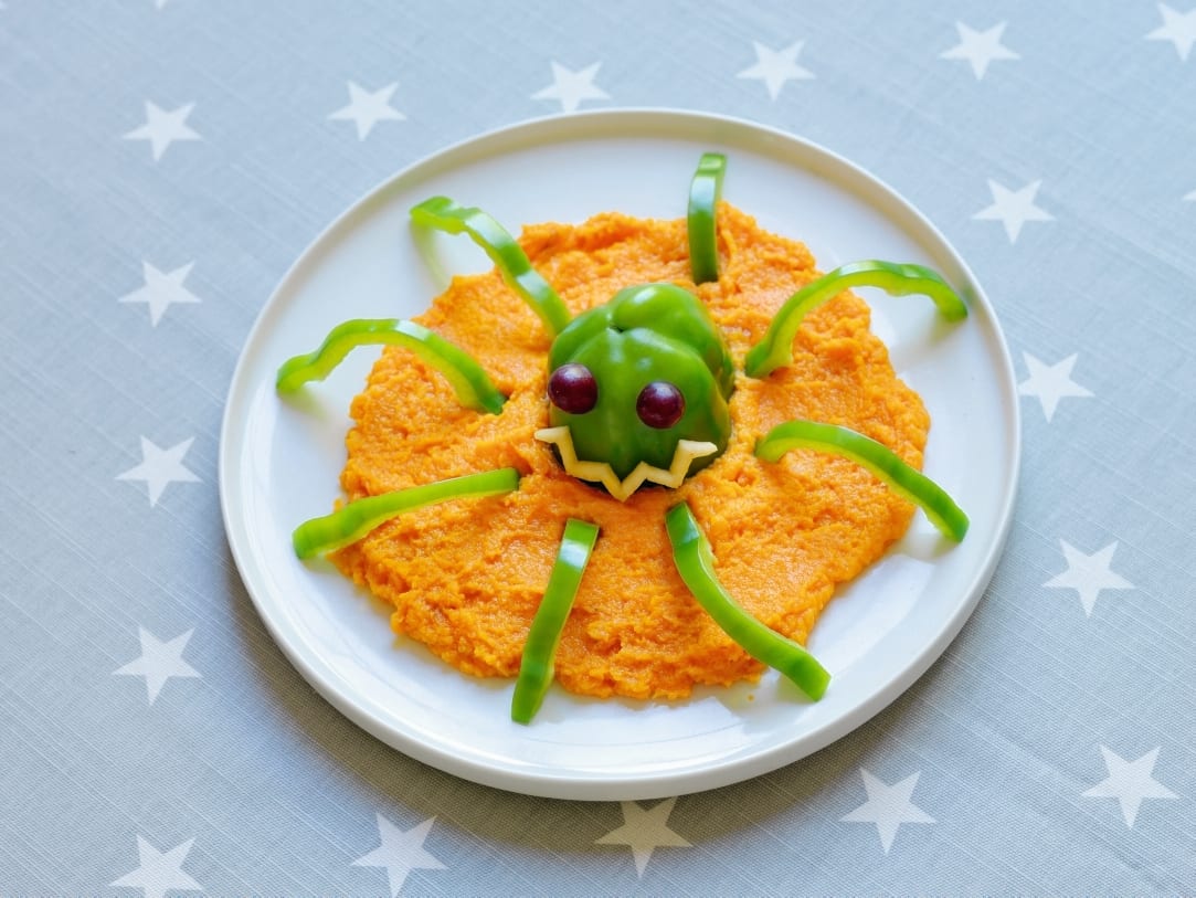 A Halloween-themed spider made from orange mashed food, a green bell pepper head, and bell pepper strip legs.