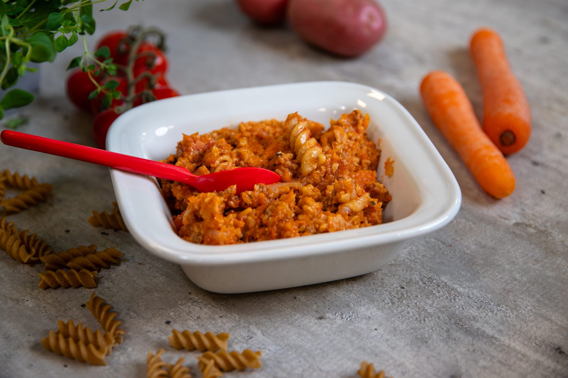 A white bowl of fusilli pasta mixed with a rich orange-red sauce and a red spoon, surrounded by raw ingredients.