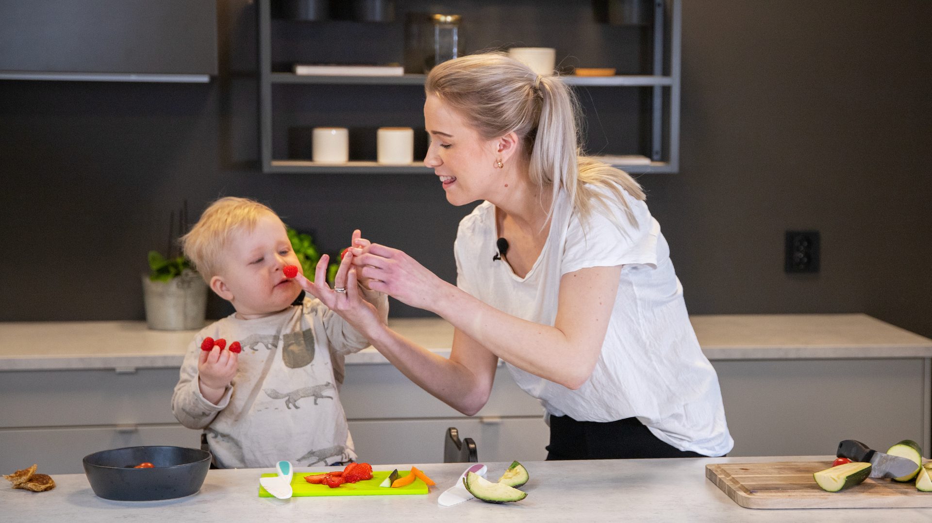 Mother and child playfully prepare fruit in a kitchen, with raspberries on their faces and hands.