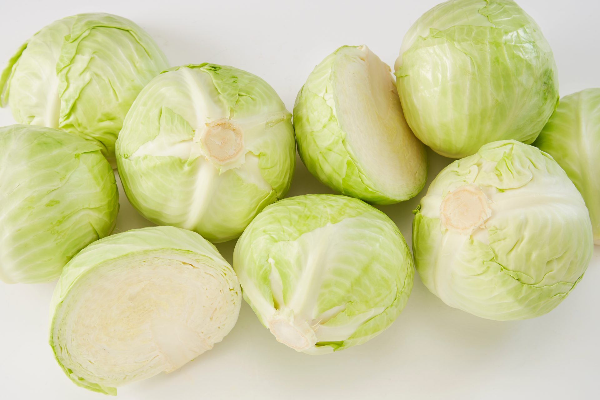 Fresh green and pale yellow cabbages, some whole and some cut, scattered on a white surface.
