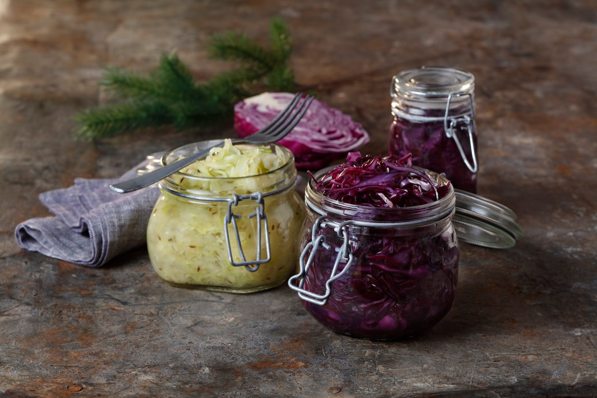 Jars of green and red sauerkraut with a fork and red cabbage slice on a rustic table.
