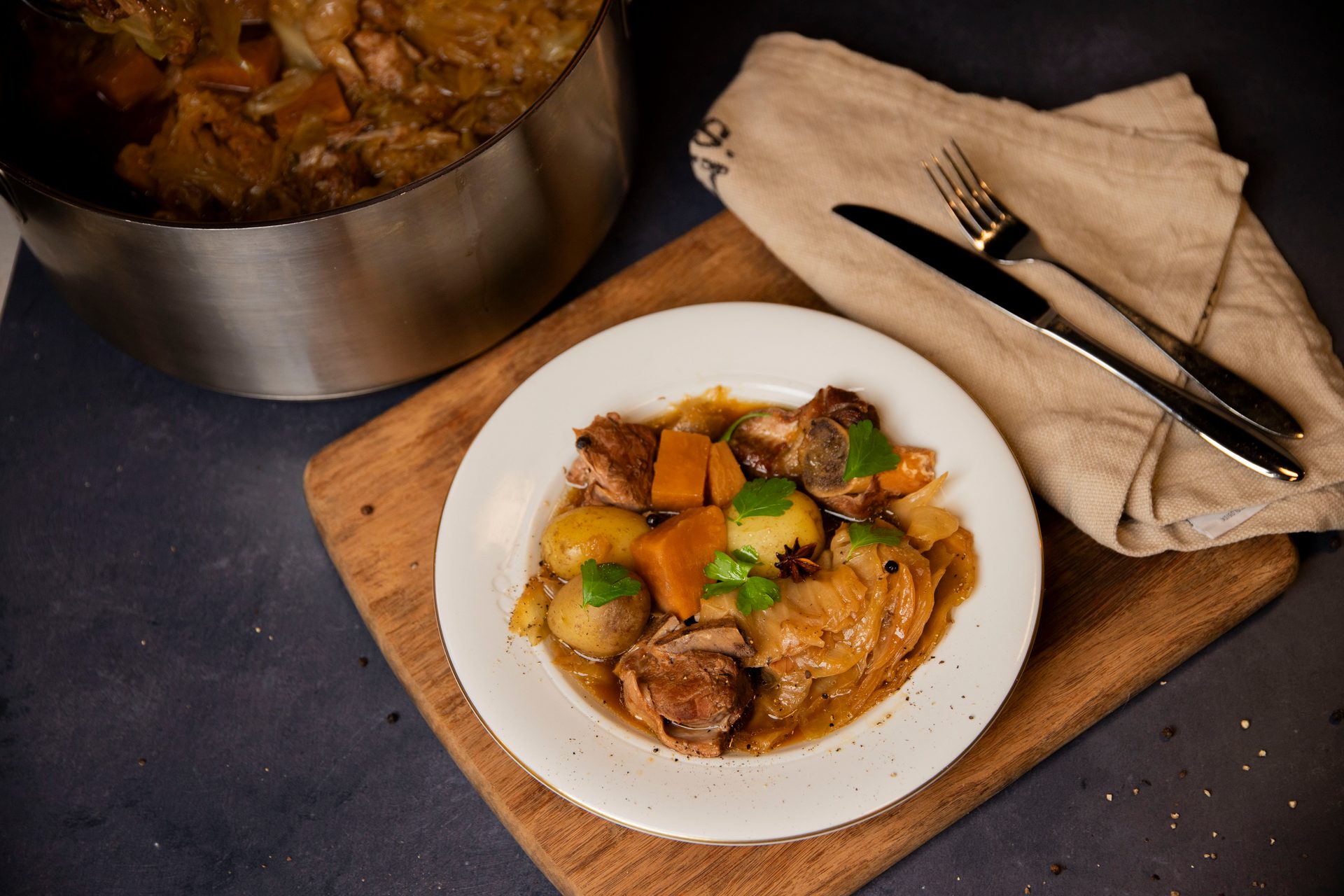 A plate of hearty meat and vegetable stew, with potatoes, cabbage, and carrots, alongside a large pot of stew, cutlery, and a napkin on a wooden board.