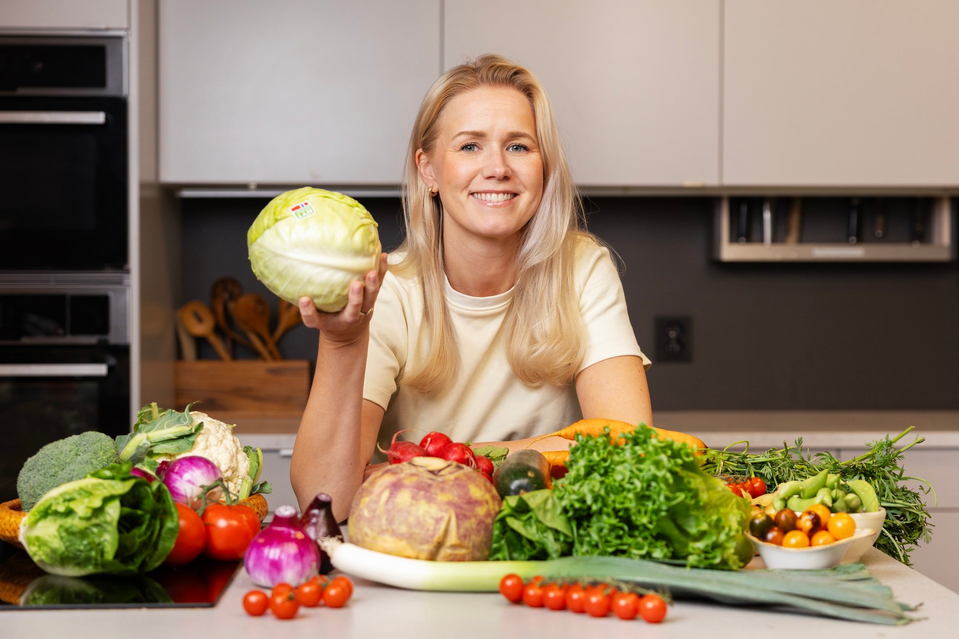 Smiling woman holding a cabbage, surrounded by a bountiful selection of fresh vegetables on a kitchen counter.