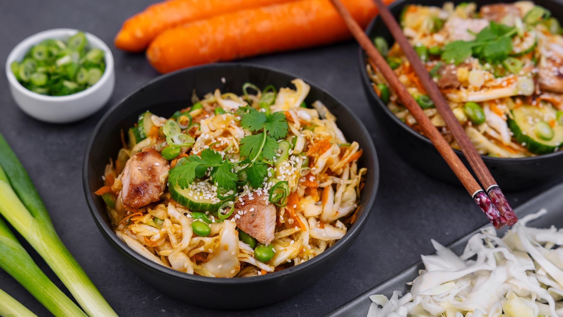 Two bowls of vibrant chicken and vegetable stir-fry with noodles, garnished with sesame seeds and fresh herbs.