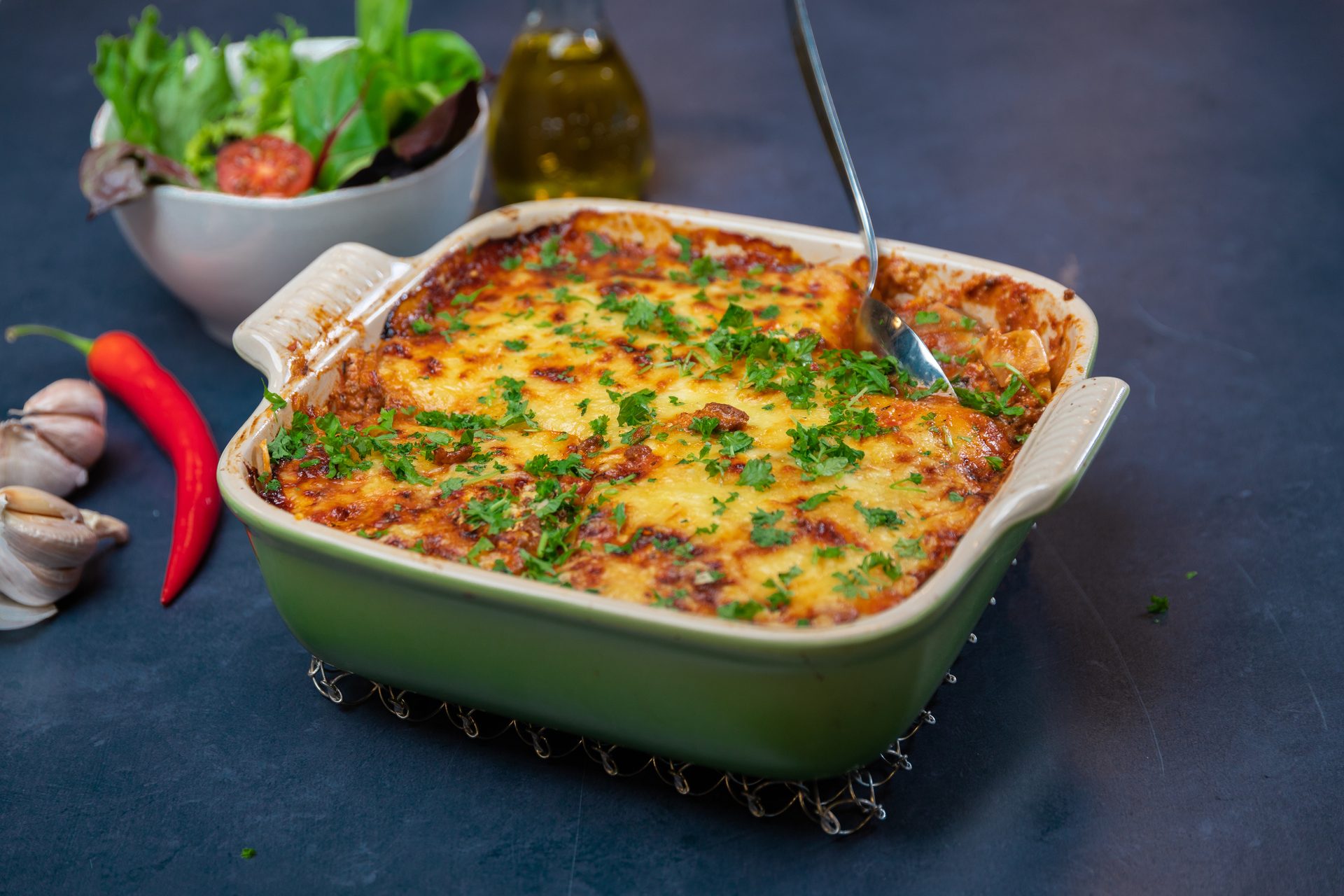 Homemade golden-baked lasagna with parsley and a spoon, served with salad, chili, and garlic.