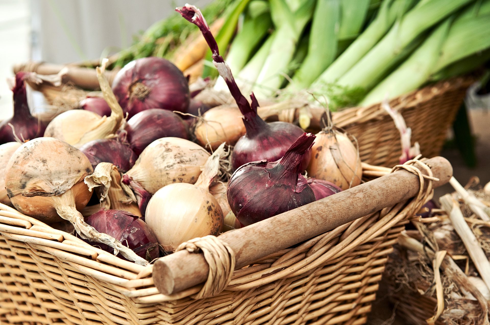 A woven basket filled with a variety of fresh red and yellow onions, with green leeks in the background.