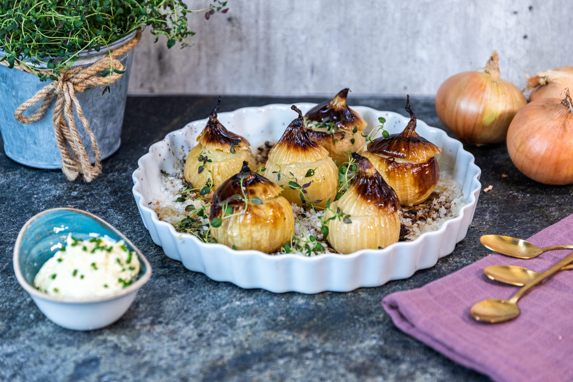 A white fluted dish holds golden-brown roasted onions with thyme, accompanied by a dip and raw onions.