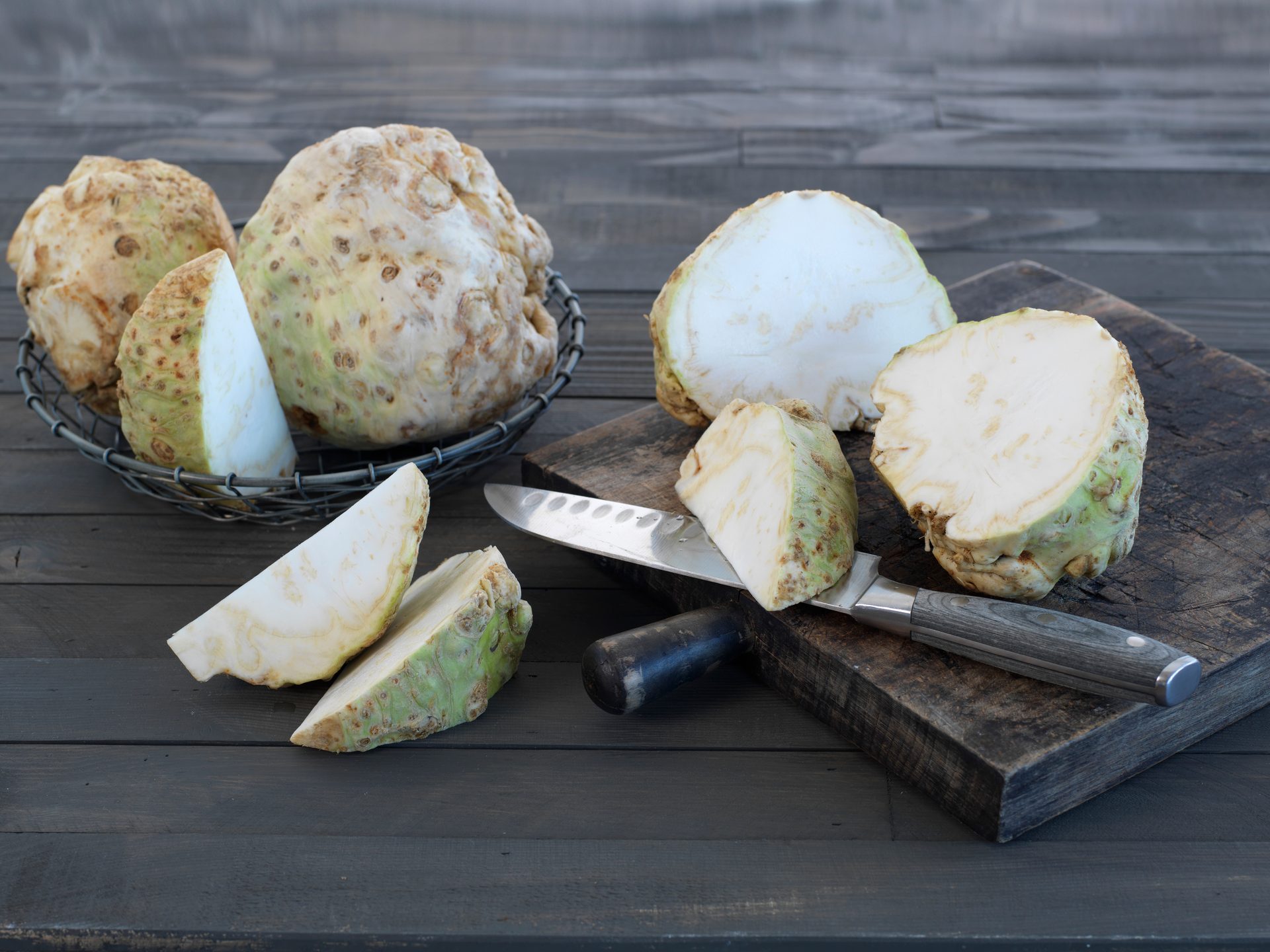 Several whole and cut celeriac roots, also known as celery root, on a dark wooden surface with a knife.