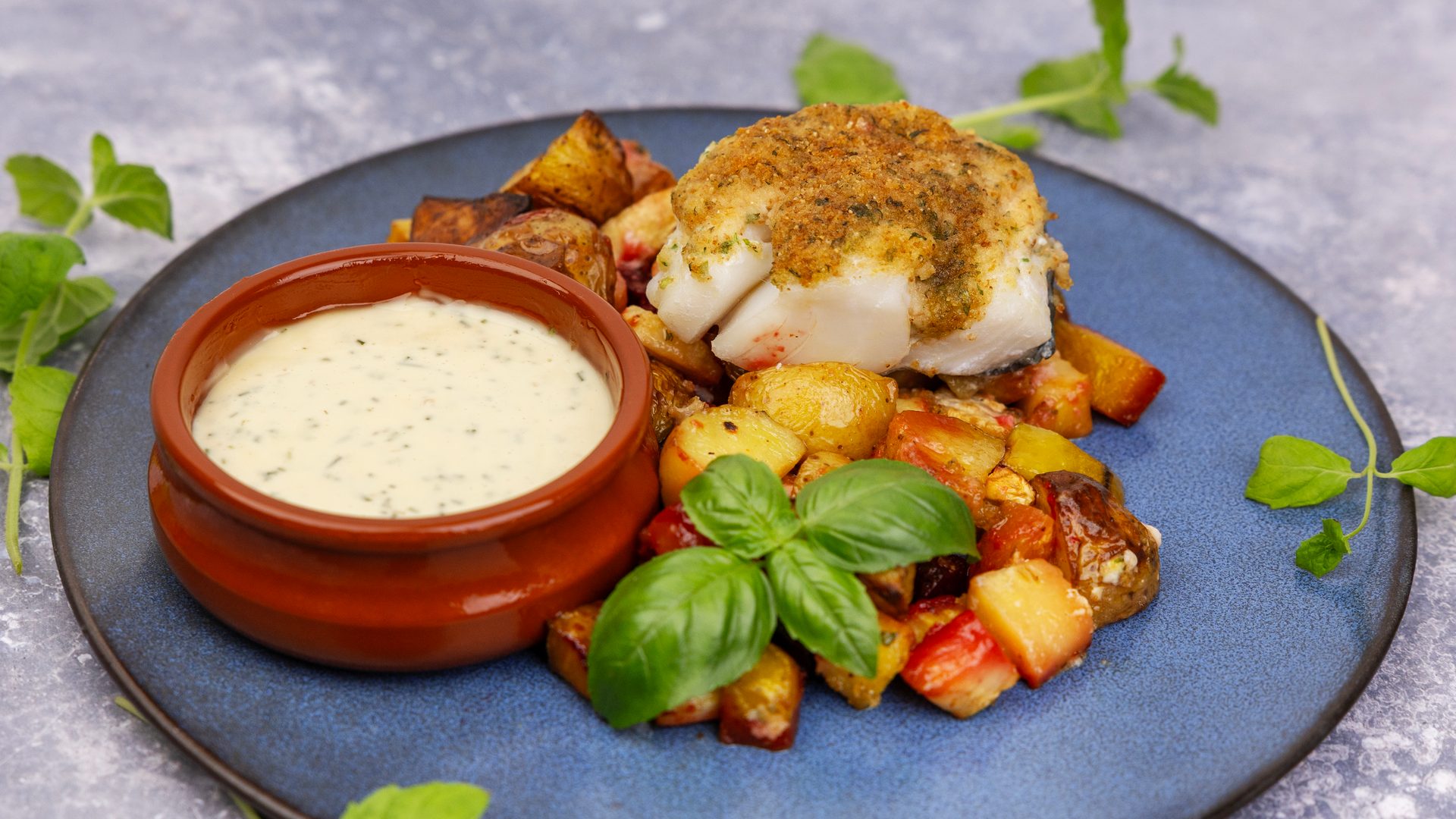 A close-up of baked cod with a crispy crust, roasted potatoes and vegetables, a dip, and fresh basil.