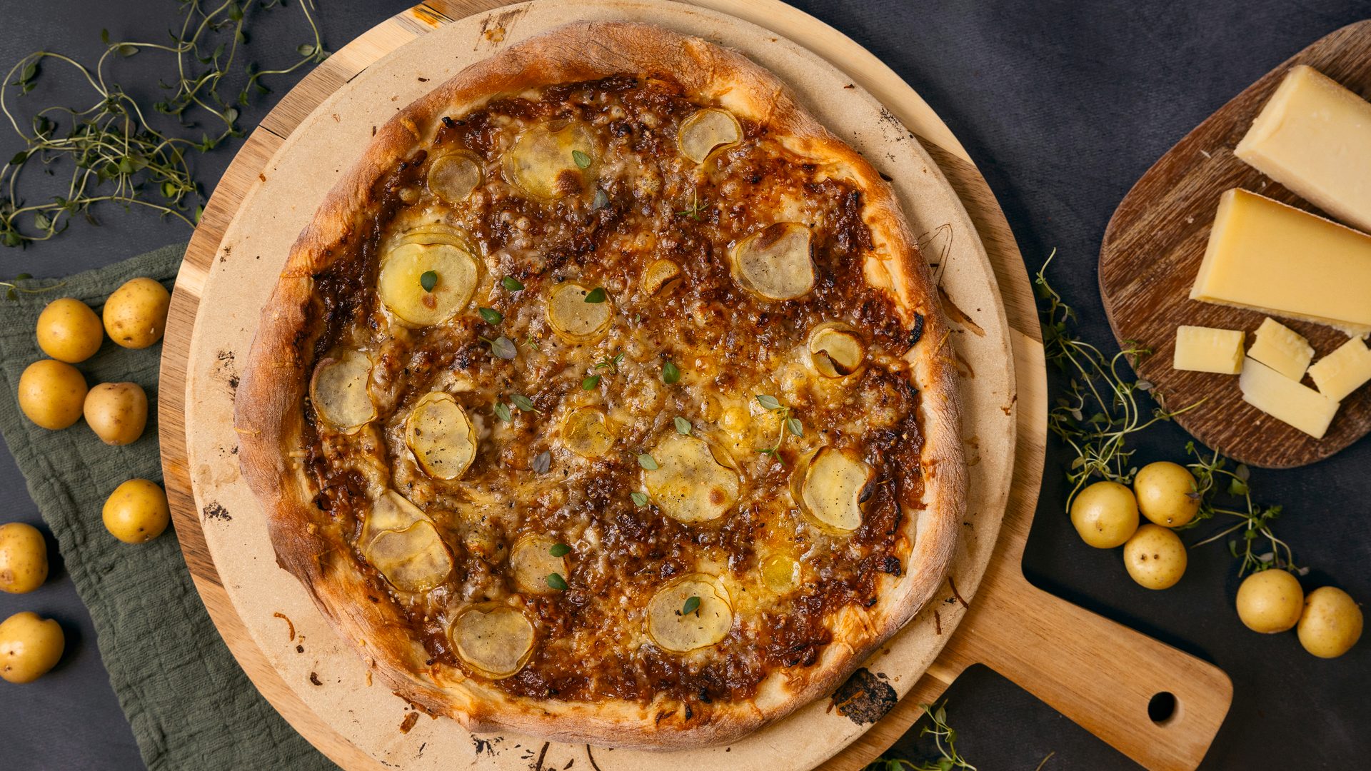 Potato and caramelized onion pizza on a peel, with cheese and raw potatoes on the side.