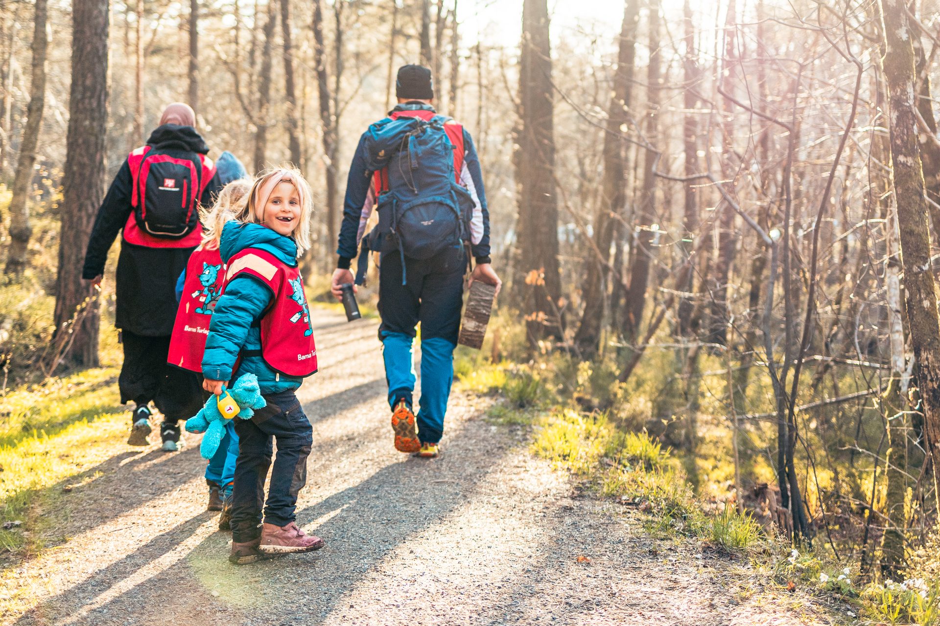 A child in a red vest smiles at the camera while hiking with family on a sunlit forest path.