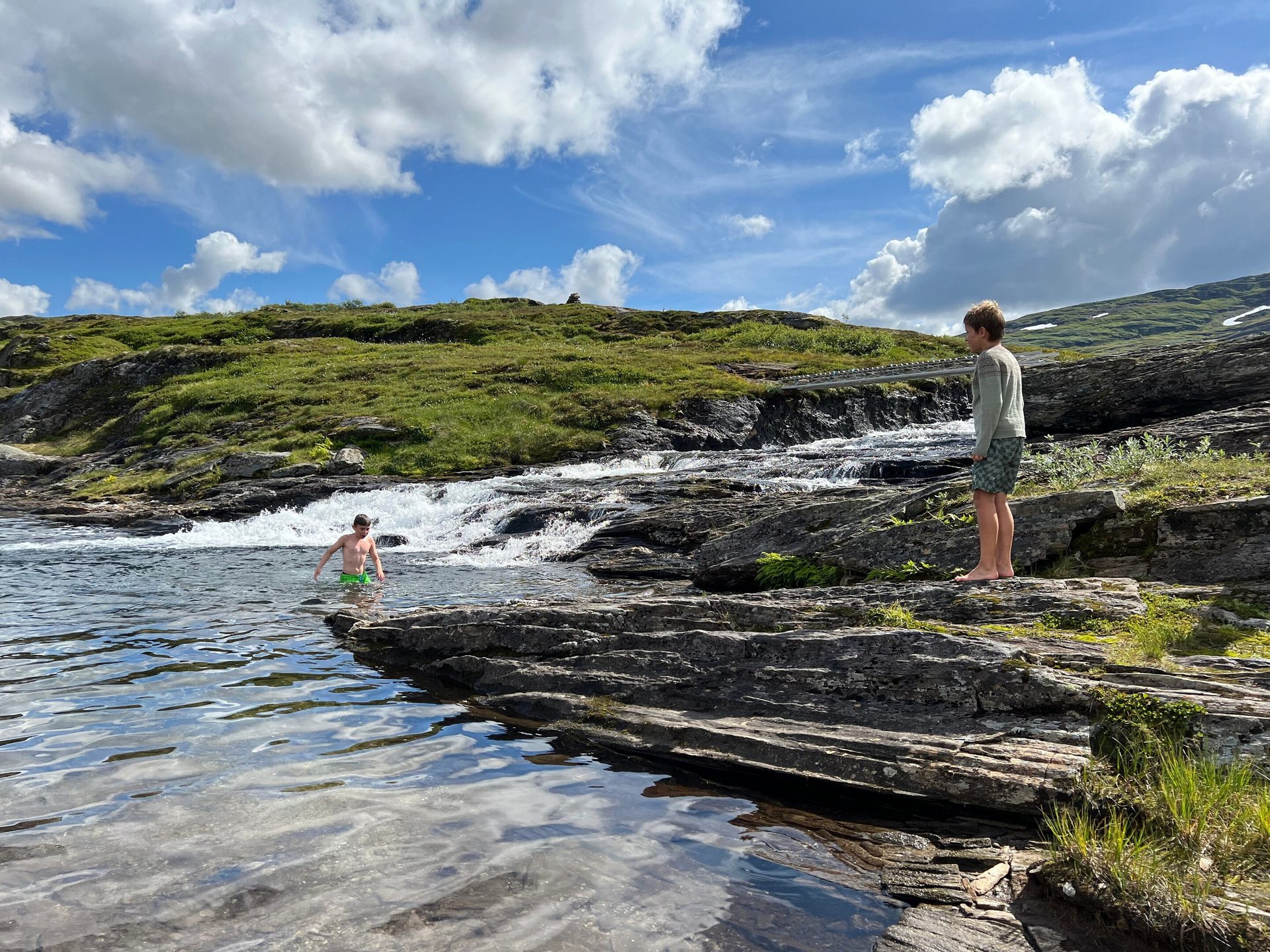 Two boys enjoy a sunny day by a rocky river with a waterfall in a green mountainous landscape.