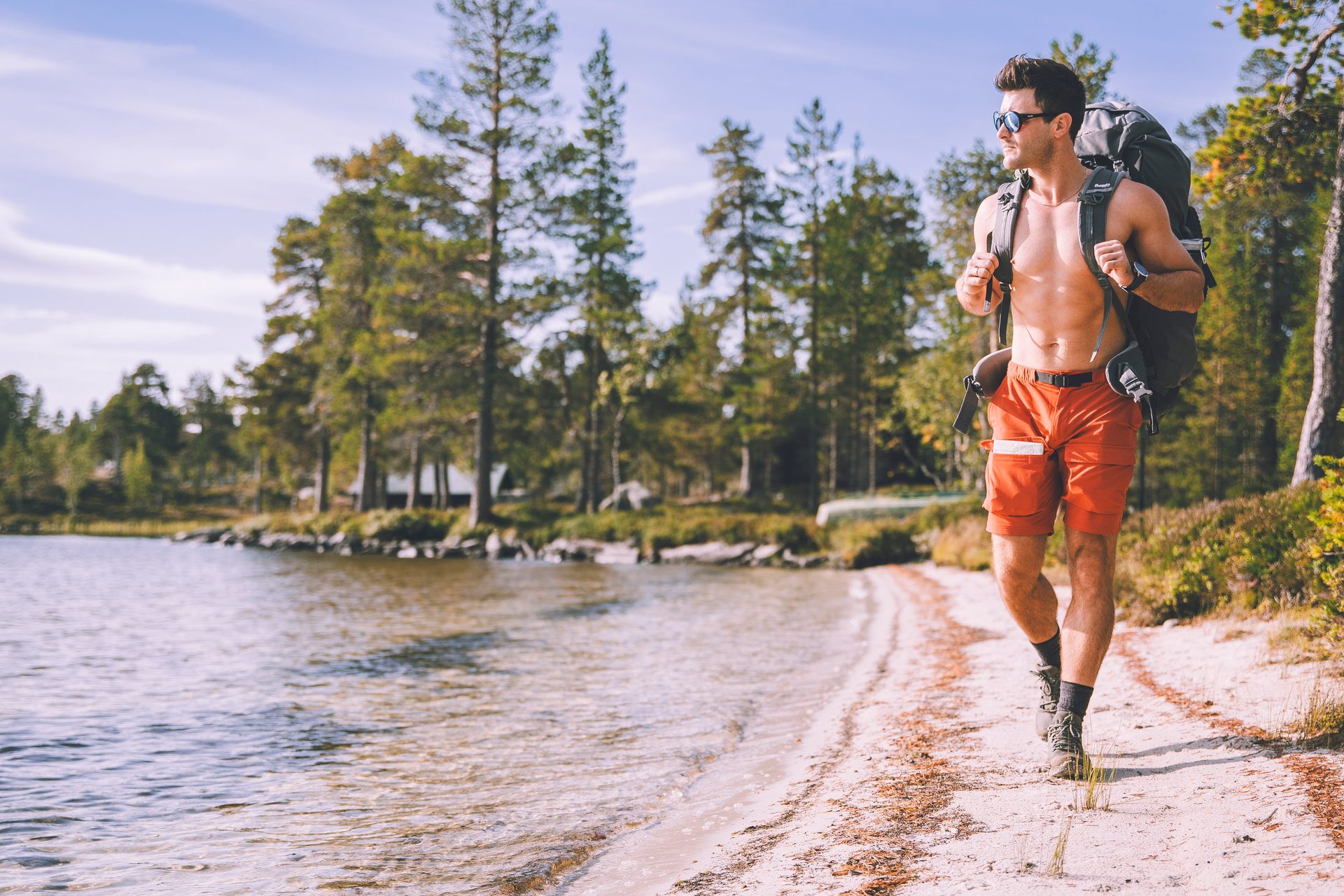 A shirtless man with a backpack hikes along a sandy lake shore lined with pine trees.