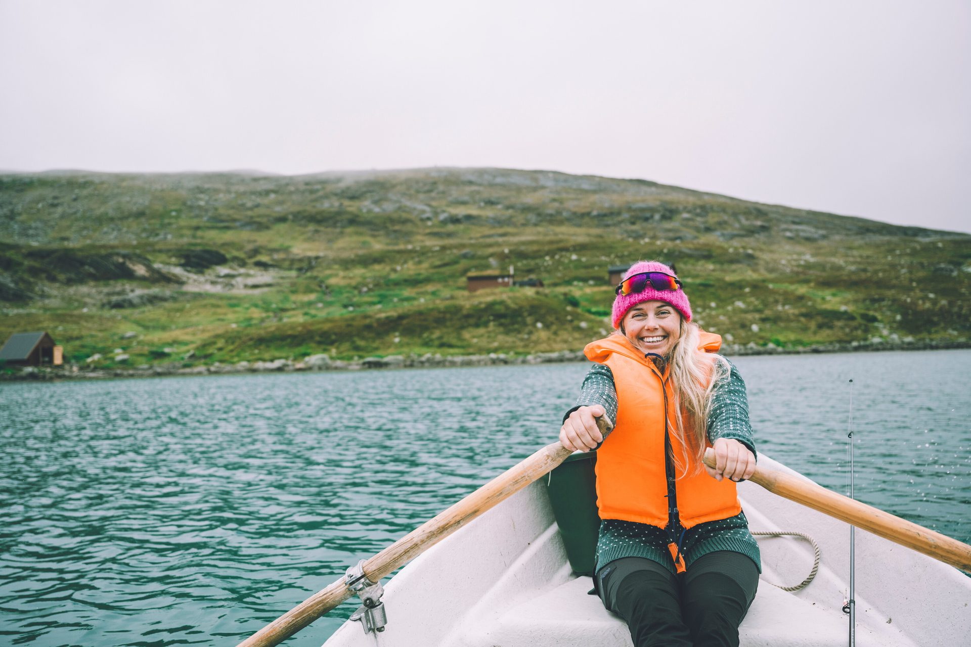 A smiling woman in an orange life vest rows a boat on a lake with green hills in the background.