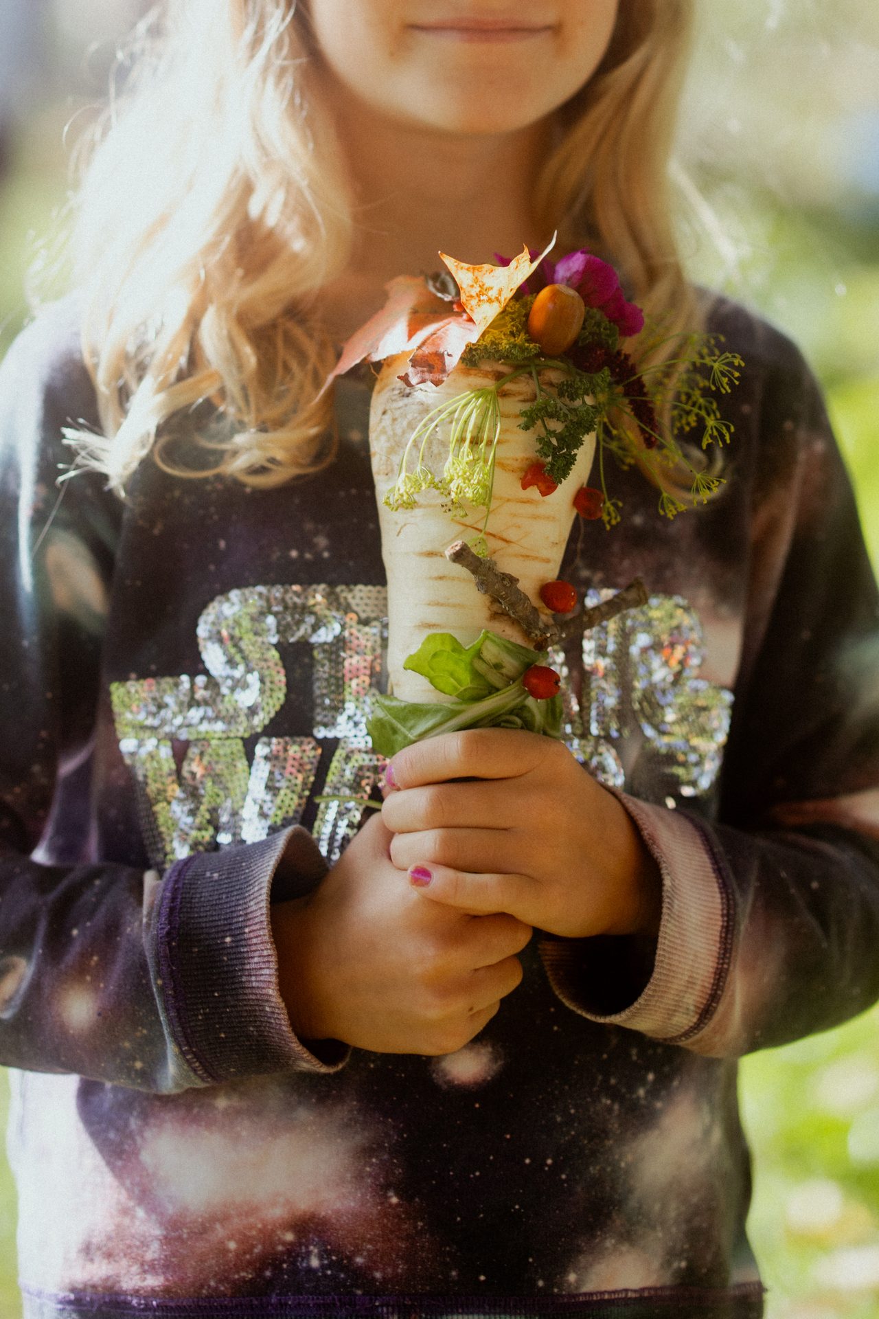 Child in a sequined Star Wars shirt holding a parsnip decorated with natural elements.