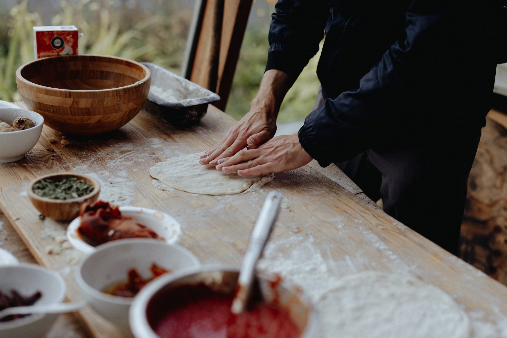 Person's hands kneading pizza dough on a floured wooden table with various ingredients nearby.