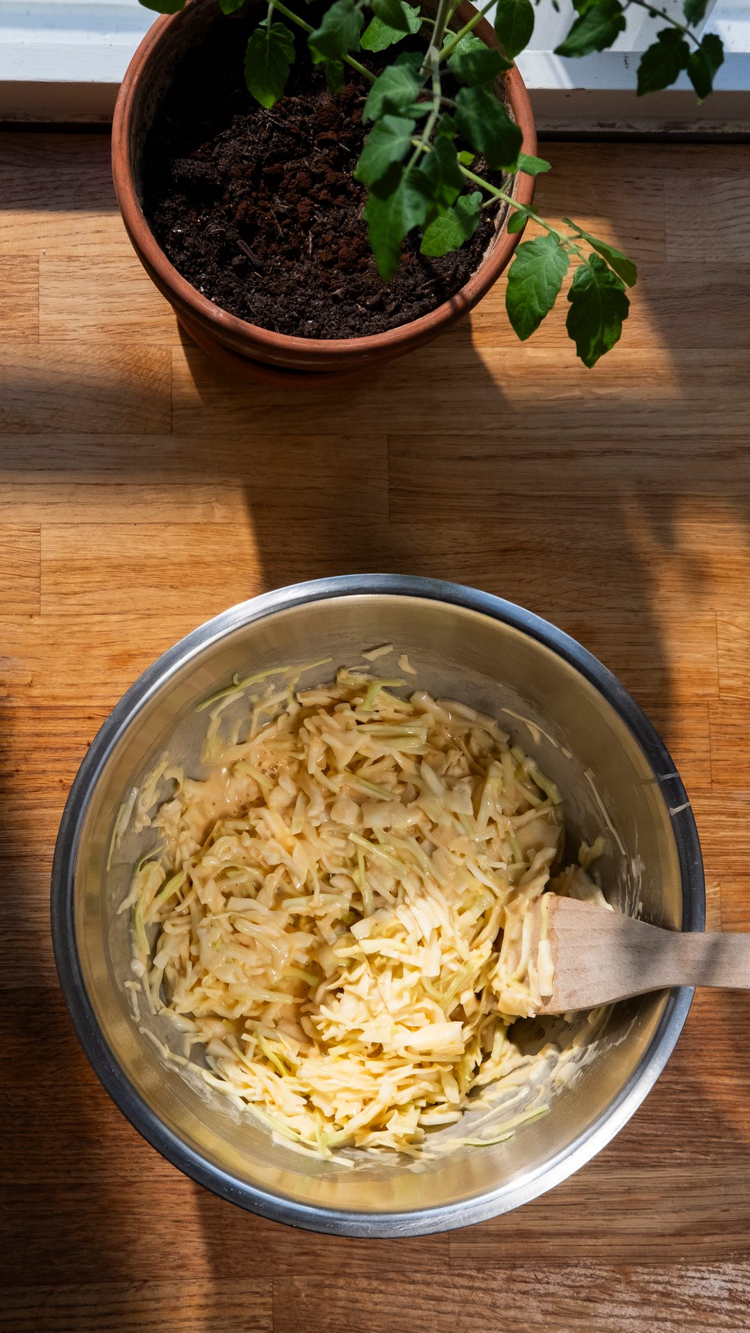 Overhead view of a bowl with shredded ingredients and a wooden spoon on a wooden counter, next to a potted plant.