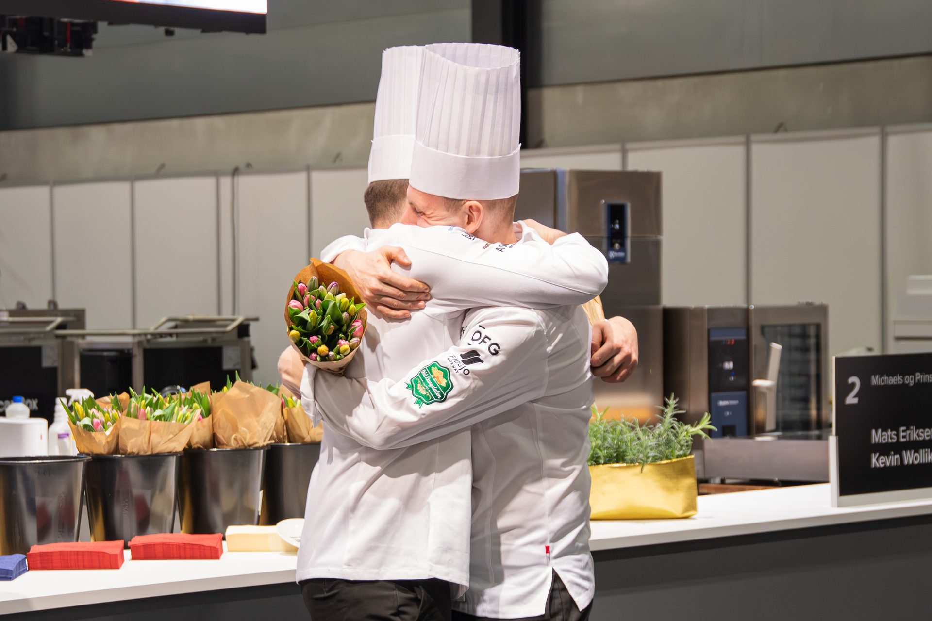 Two chefs in white uniforms and tall hats embrace, one holding a bouquet of tulips, celebrating a victory.