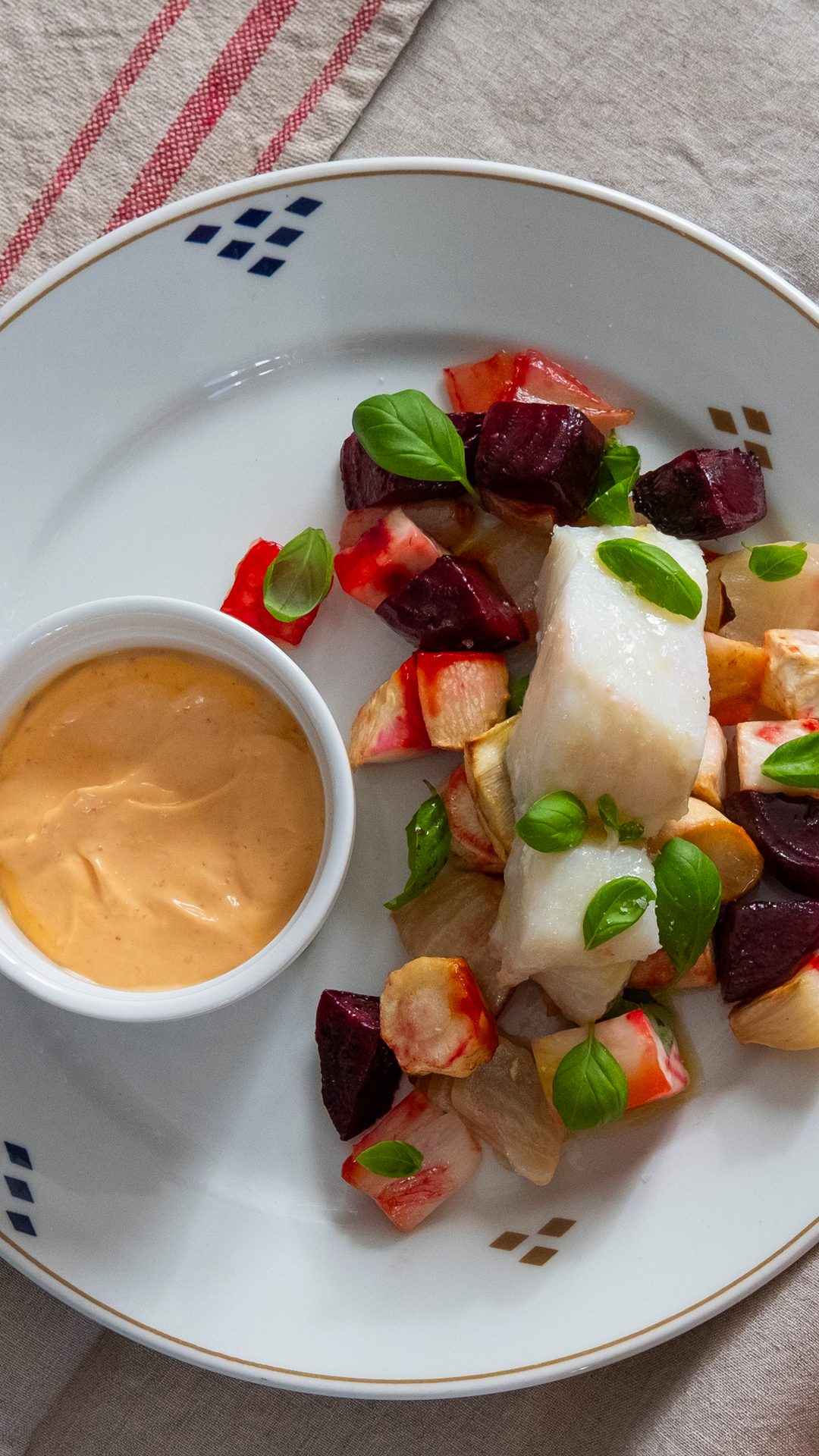 A white plate with a piece of flaky white fish, roasted root vegetables, fresh basil, and a side of orange sauce.