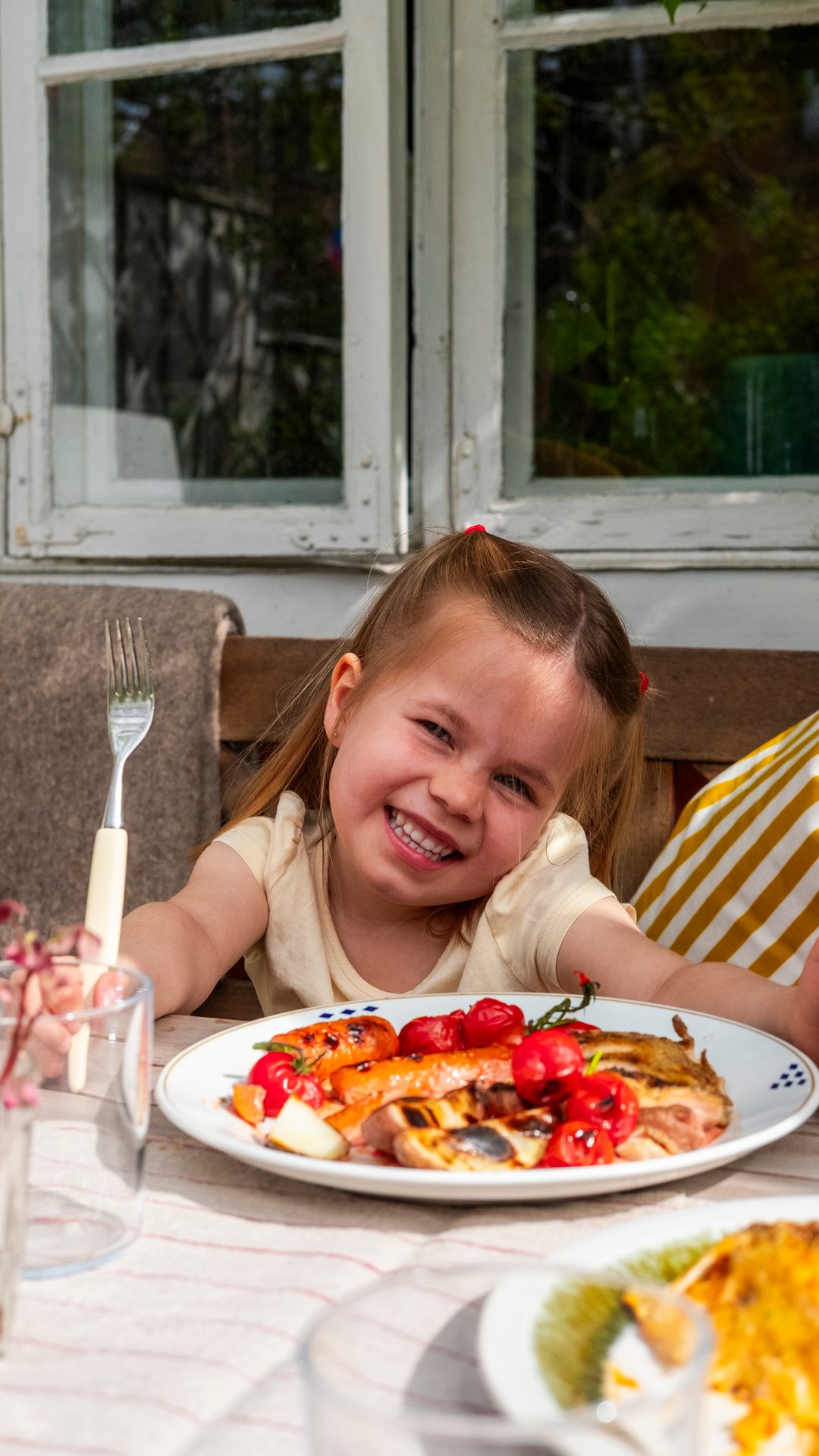 A happy young girl smiling while sitting at an outdoor table with a plate of grilled food.