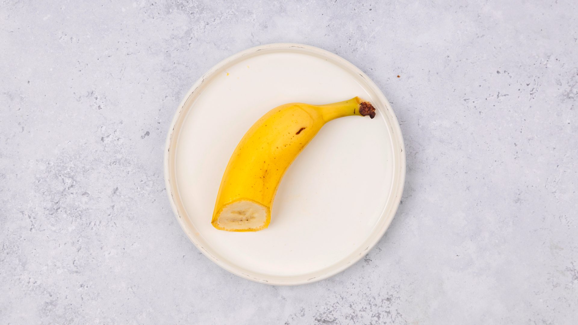 Half a yellow banana on a white plate against a grey, textured background.