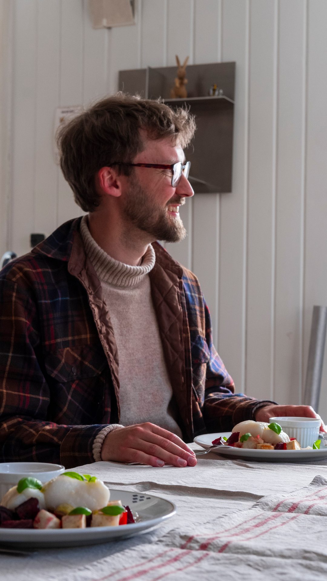 A man in glasses and a plaid shirt smiles while sitting at a table with two plates of food.