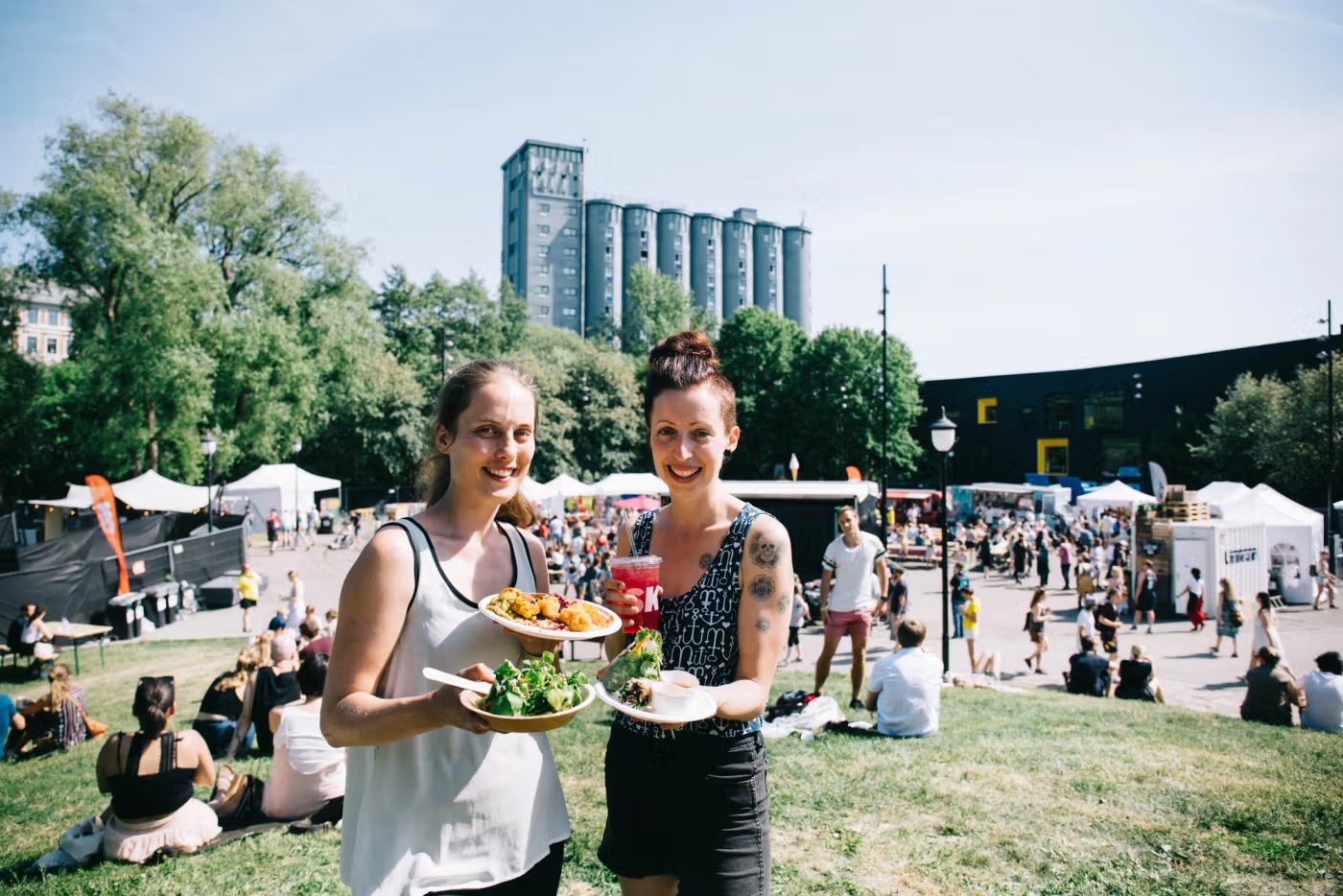 Two smiling women with plates of food and a drink at an outdoor market on a sunny day.