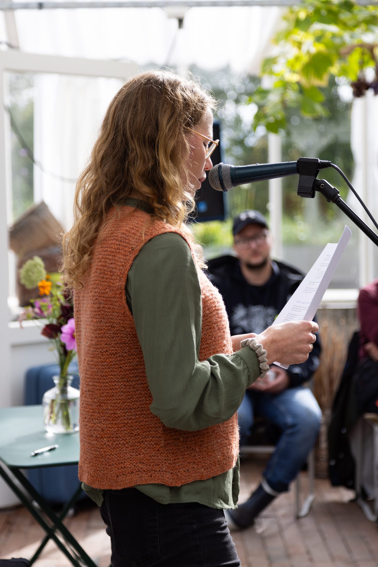 A woman with curly hair and glasses reads from papers into a microphone, wearing an orange vest and green shirt.