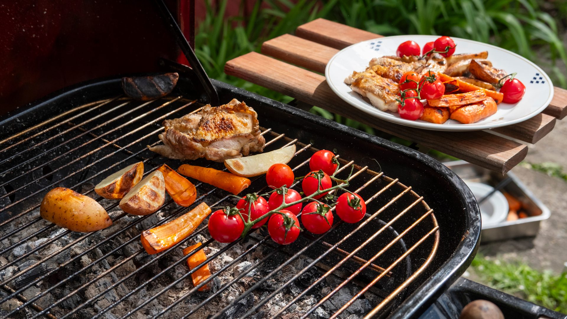 Grilling chicken, potatoes, carrots, and tomatoes on a barbecue, with a plate of cooked food nearby.