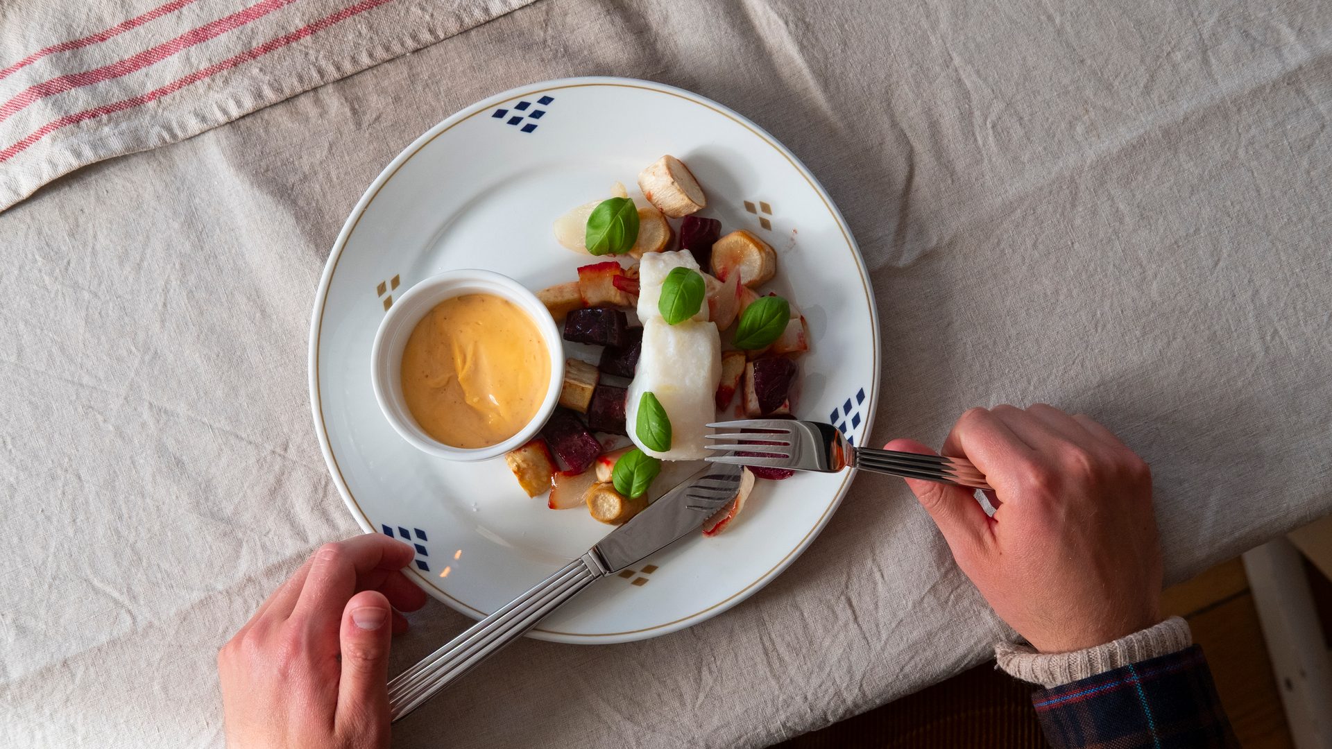 Person cutting a white fish dish with roasted root vegetables, basil, and a side of orange sauce on a white plate.
