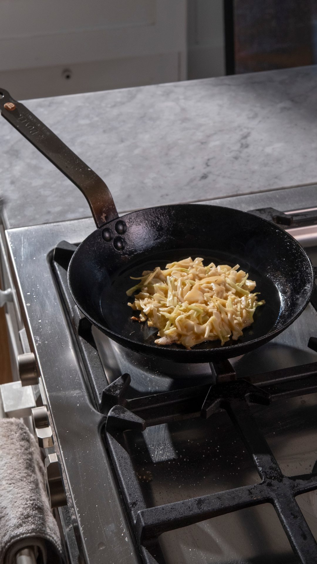 A pan with shredded vegetables cooking on a gas stove burner with a marble countertop.