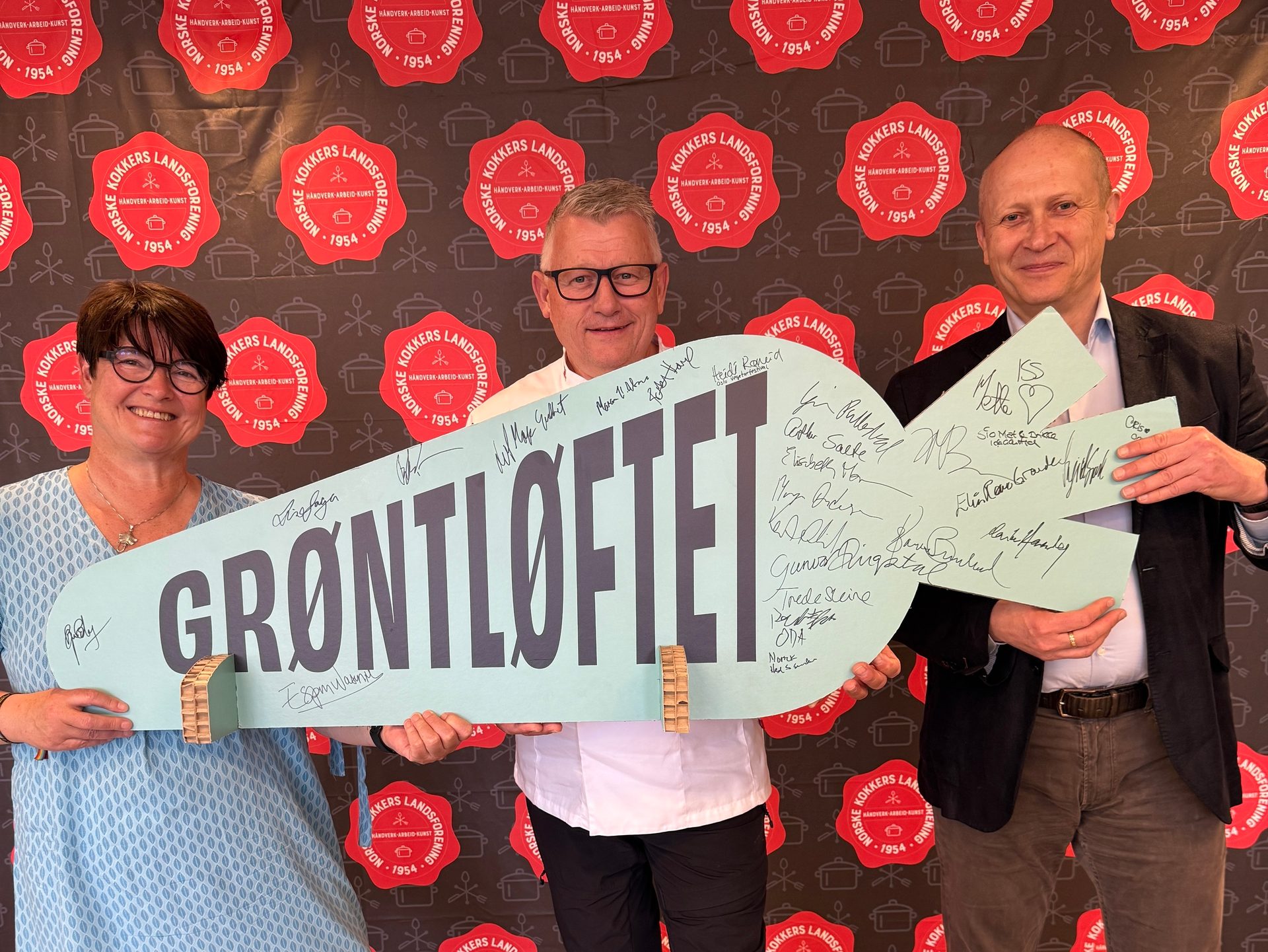 Three people hold a large, key-shaped "GRØNTLØFTET" sign with signatures, in front of a chefs' association backdrop.