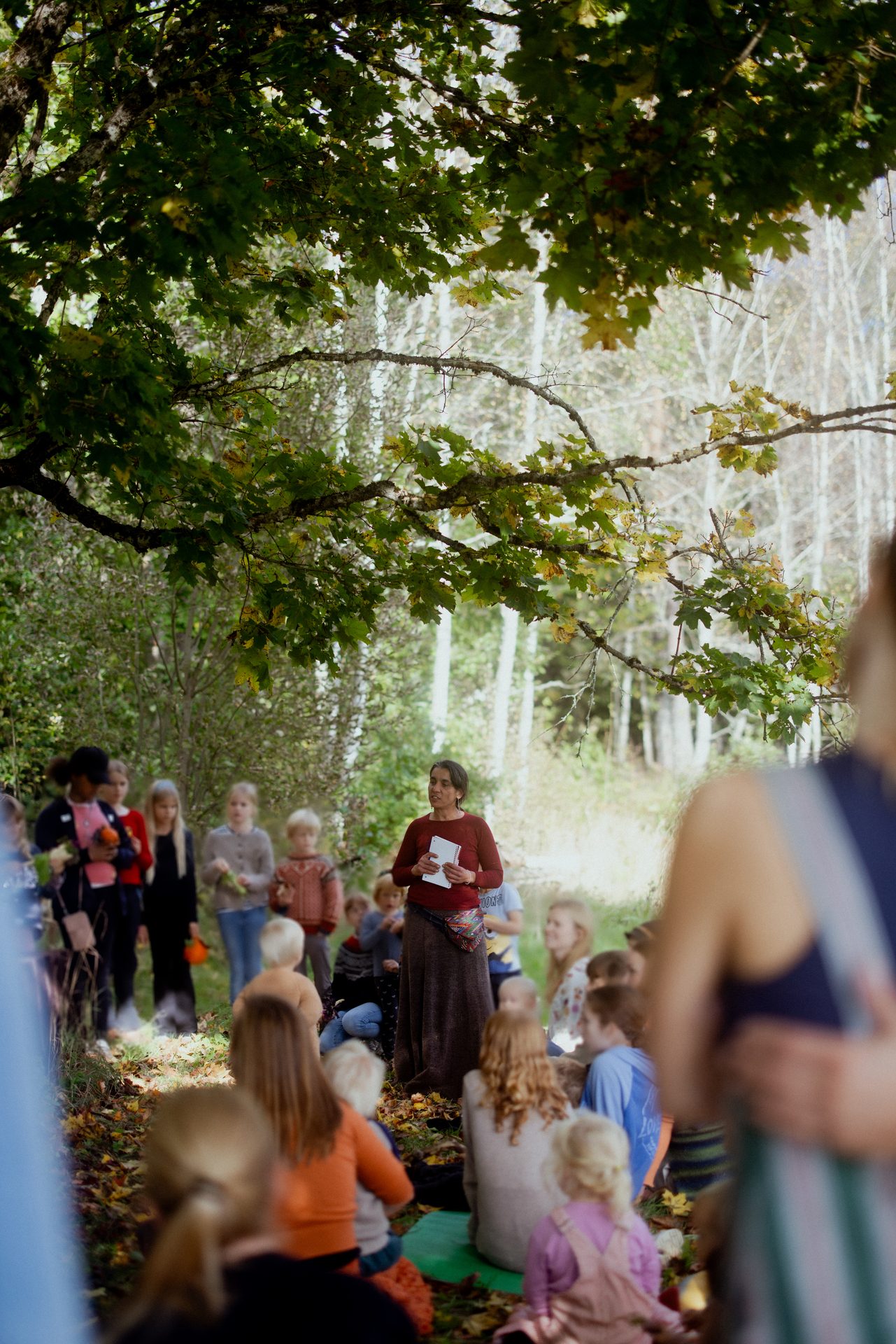 A woman addresses a group of children and adults in an outdoor nature setting, surrounded by trees.