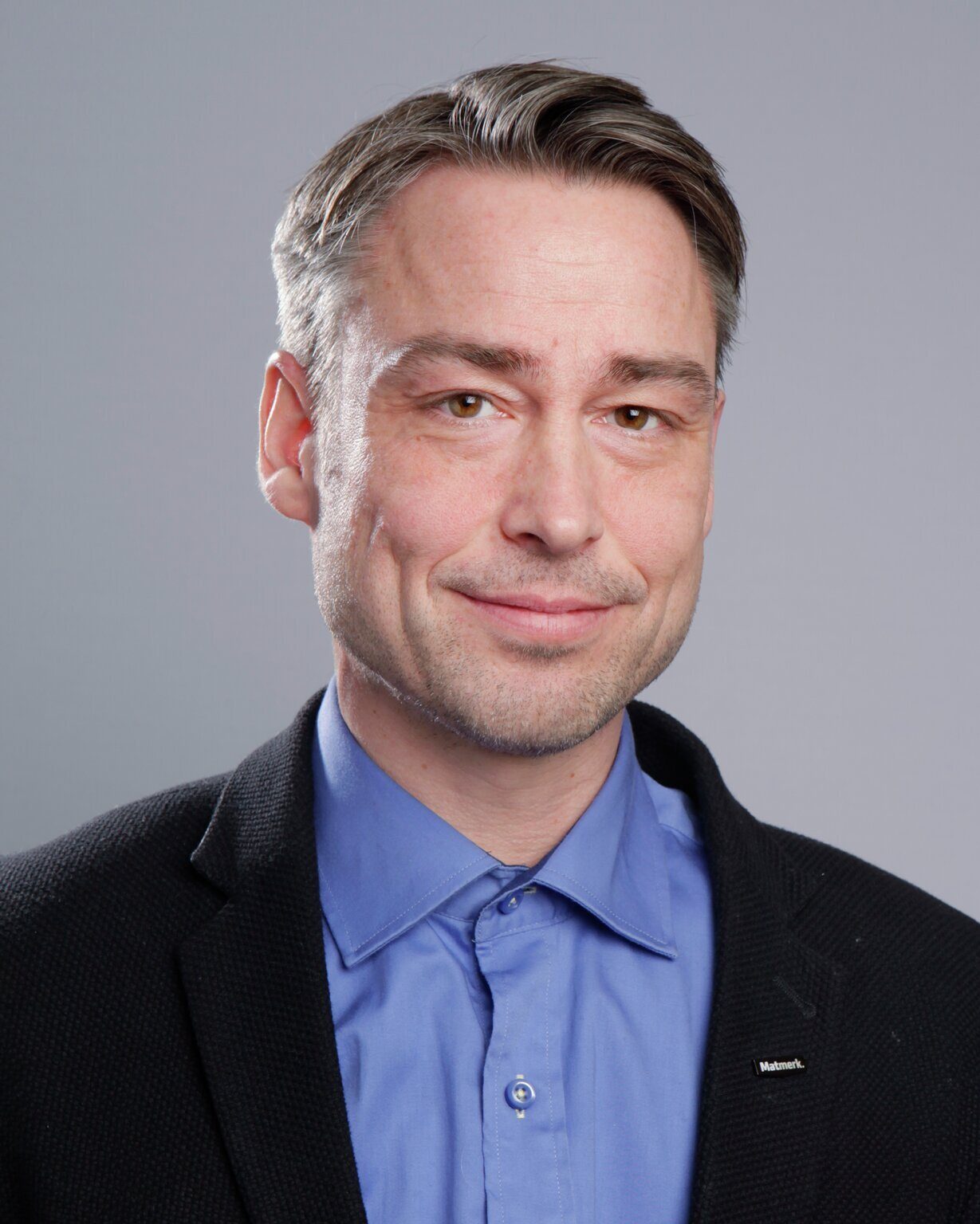Headshot of a smiling man with short graying hair, wearing a blue shirt and black jacket. He looks friendly.
