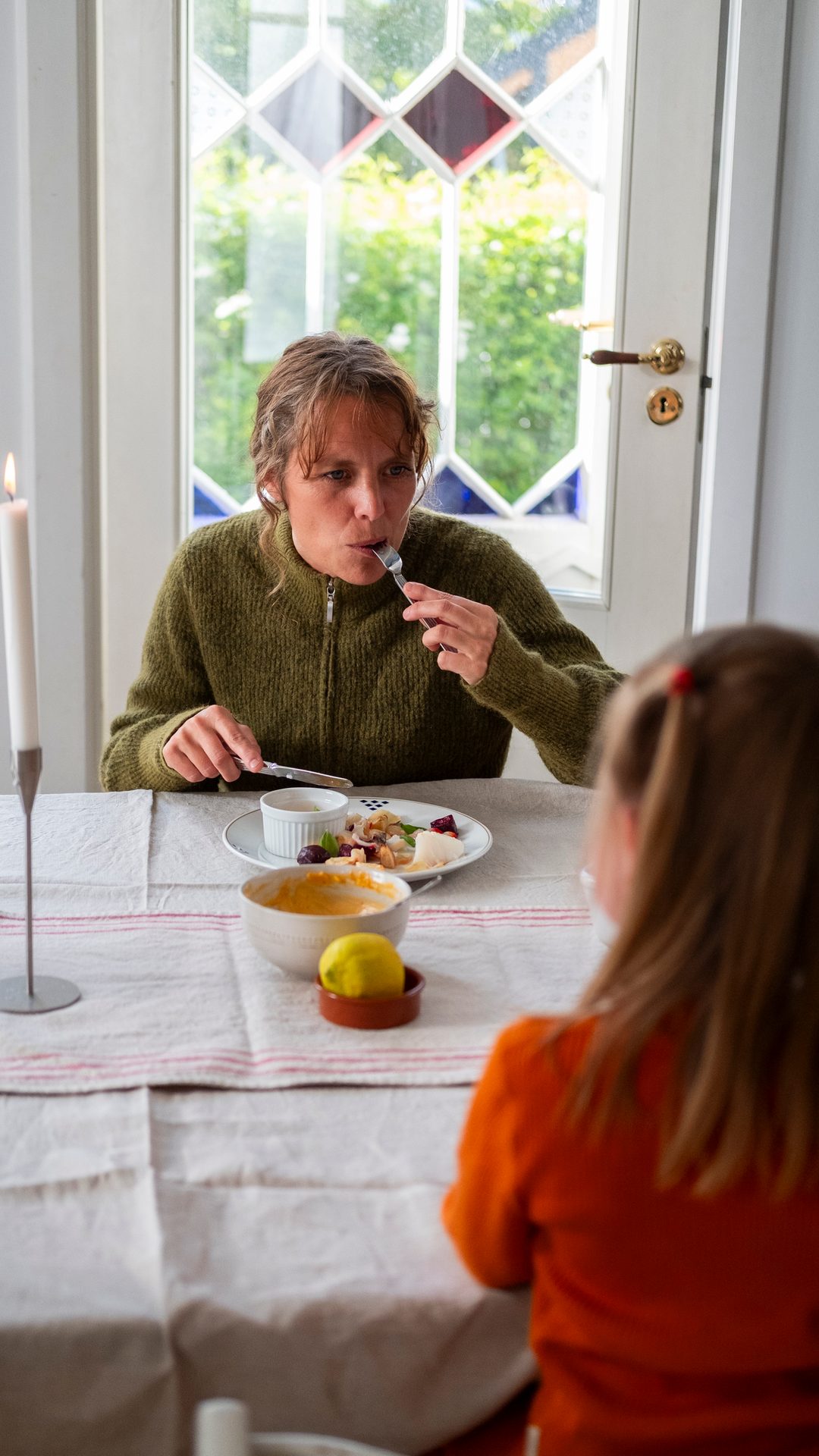 Woman in green sweater eating at a table with a child in the foreground, by a window.