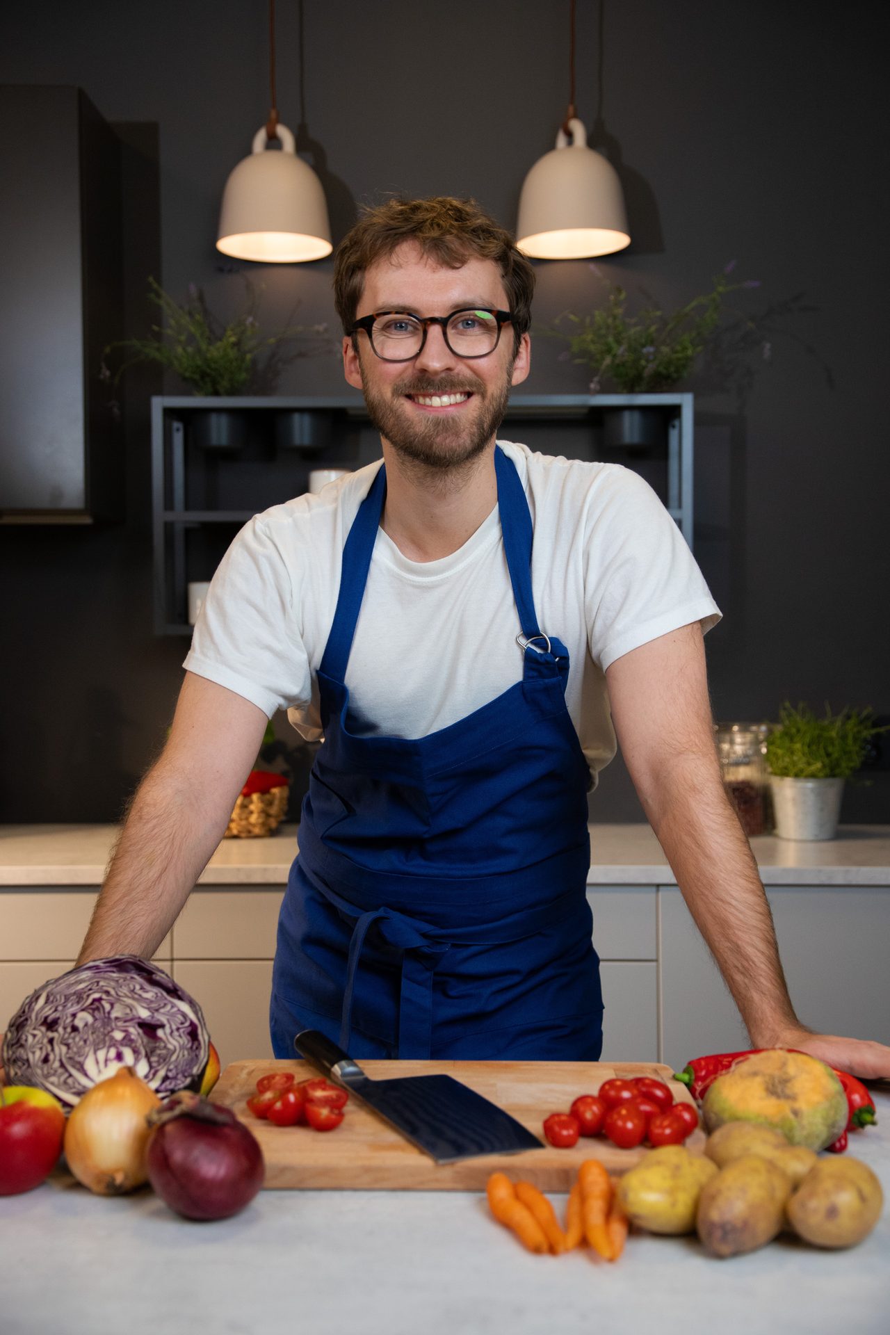 Smiling chef in blue apron in modern kitchen, surrounded by fresh vegetables on counter.