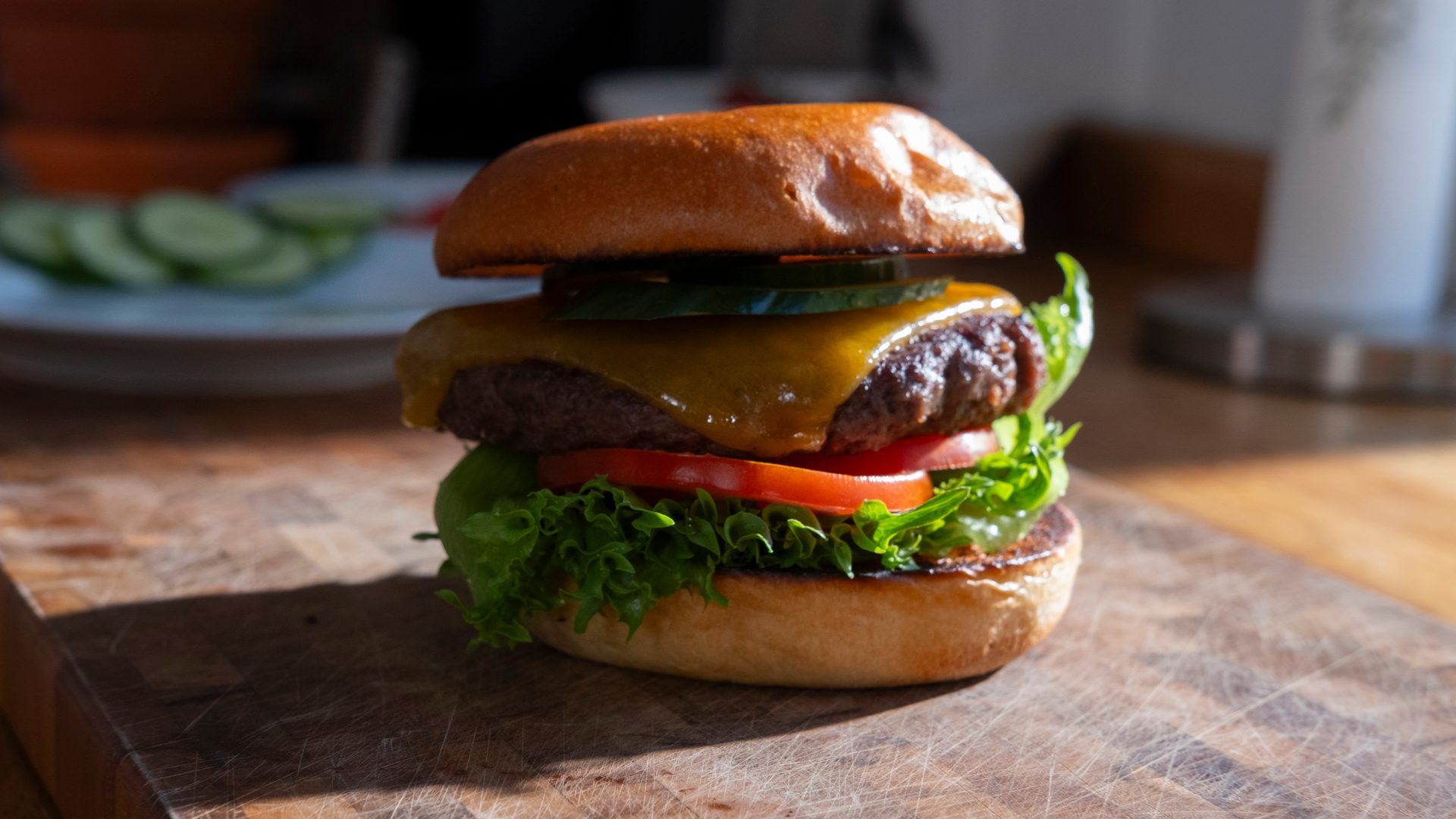 A delicious cheeseburger with lettuce, tomato, cucumber, and melted cheese on a bun on a cutting board.