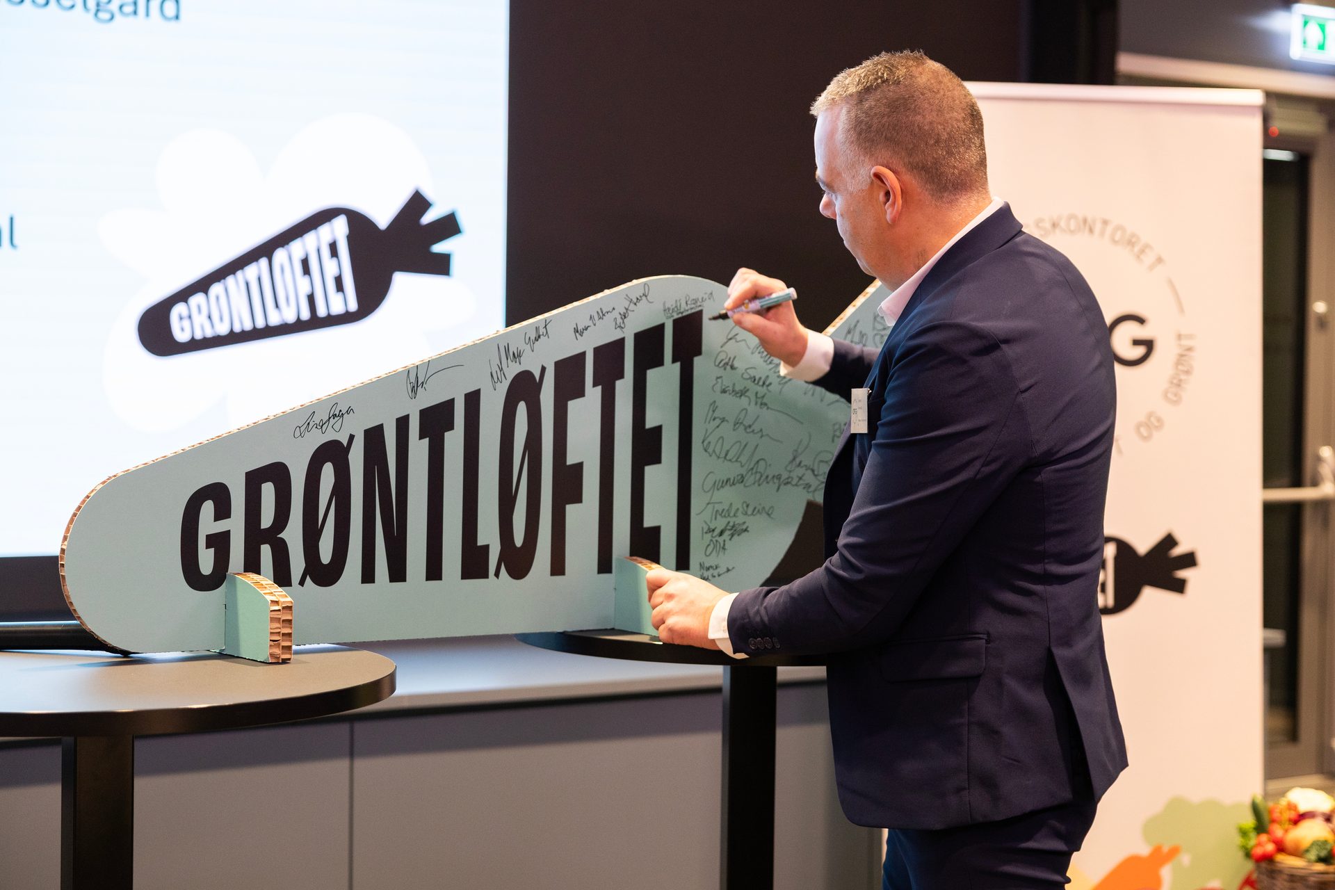 A man in a suit signs a large "GRØNTLØFTET" display board. A logo is on a screen in the background.