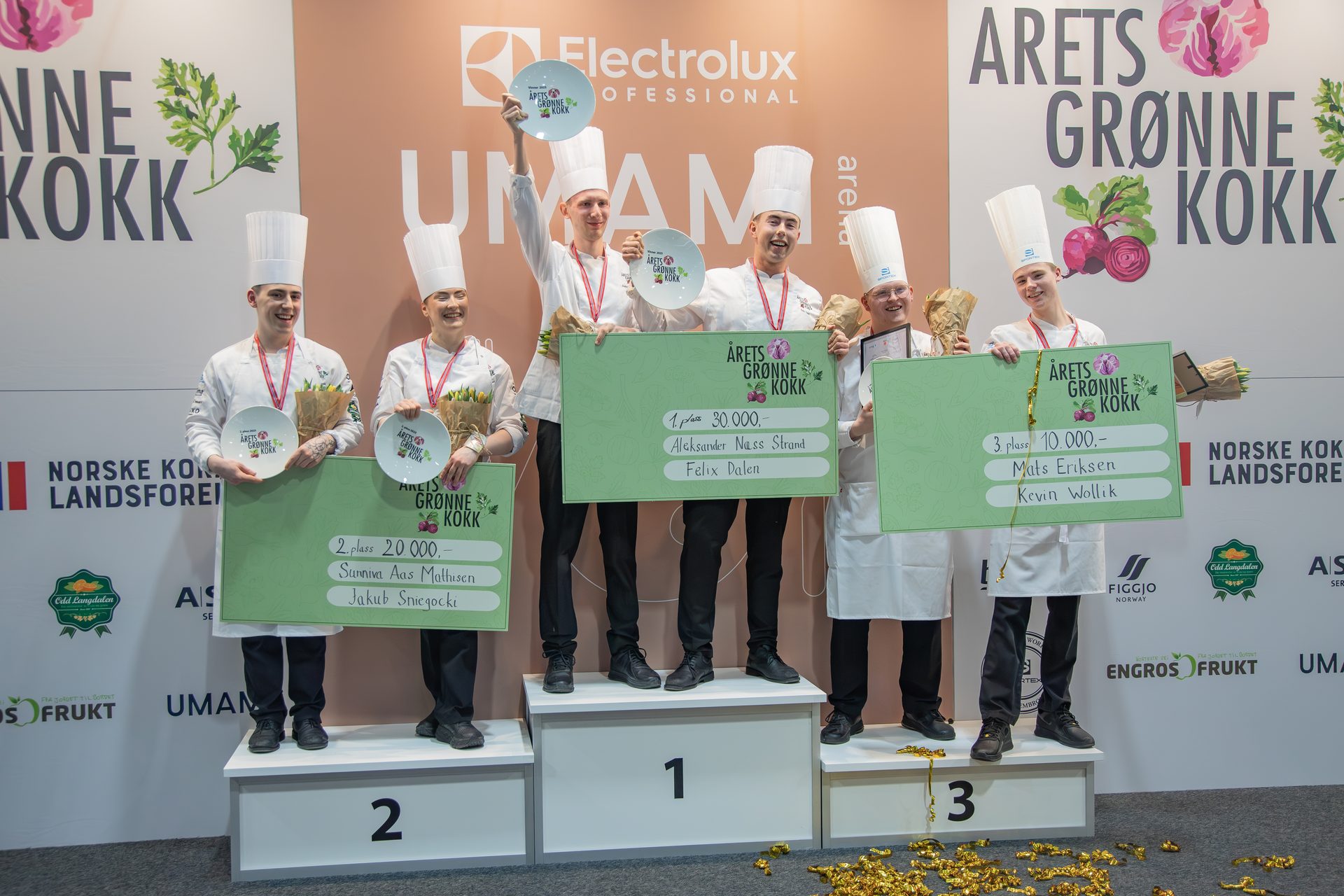 Six young chefs smiling on a podium, holding their awards and large prize checks from a cooking competition.